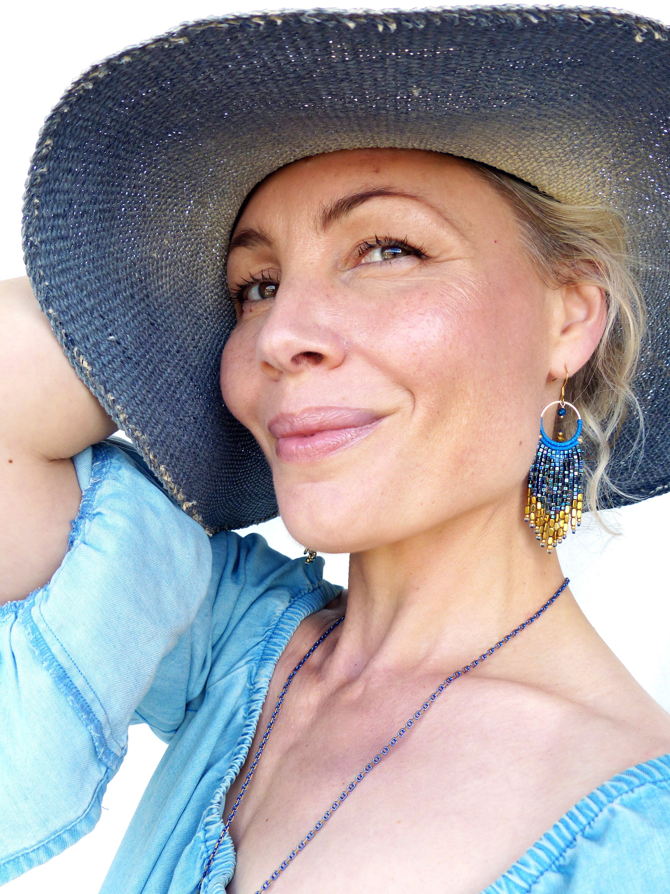 Peut inclure: Une femme portant une chemise en jean bleu et un chapeau de paille à larges bords. Elle porte un collier de perles et des boucles d'oreilles avec des perles bleues et dorées.