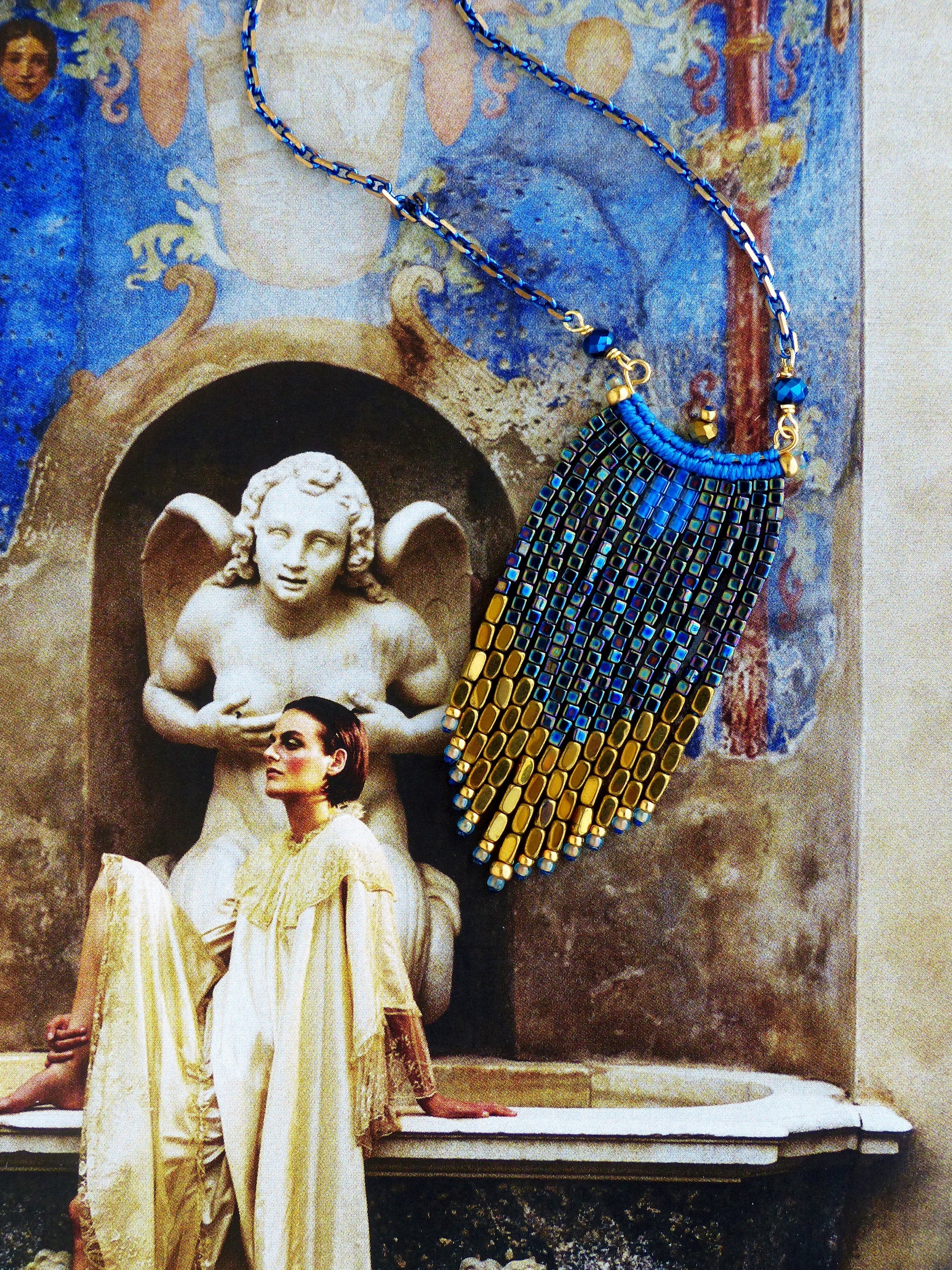 Peut inclure: Un collier de perles dorées et bleues avec un design en frange. Le collier est suspendu devant une fontaine en pierre avec une statue d'ange.