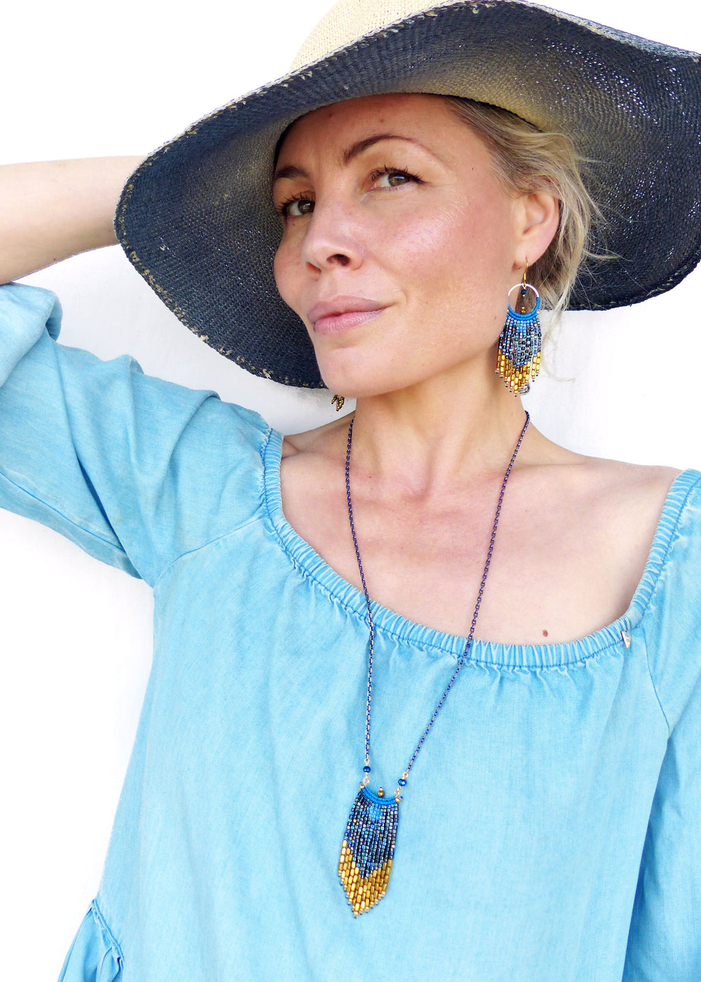 Peut inclure: Une femme portant un haut en jean bleu décolleté, un chapeau de paille à larges bords avec un bandeau bleu, et un collier et des boucles d'oreilles en perles dorées et bleues.
