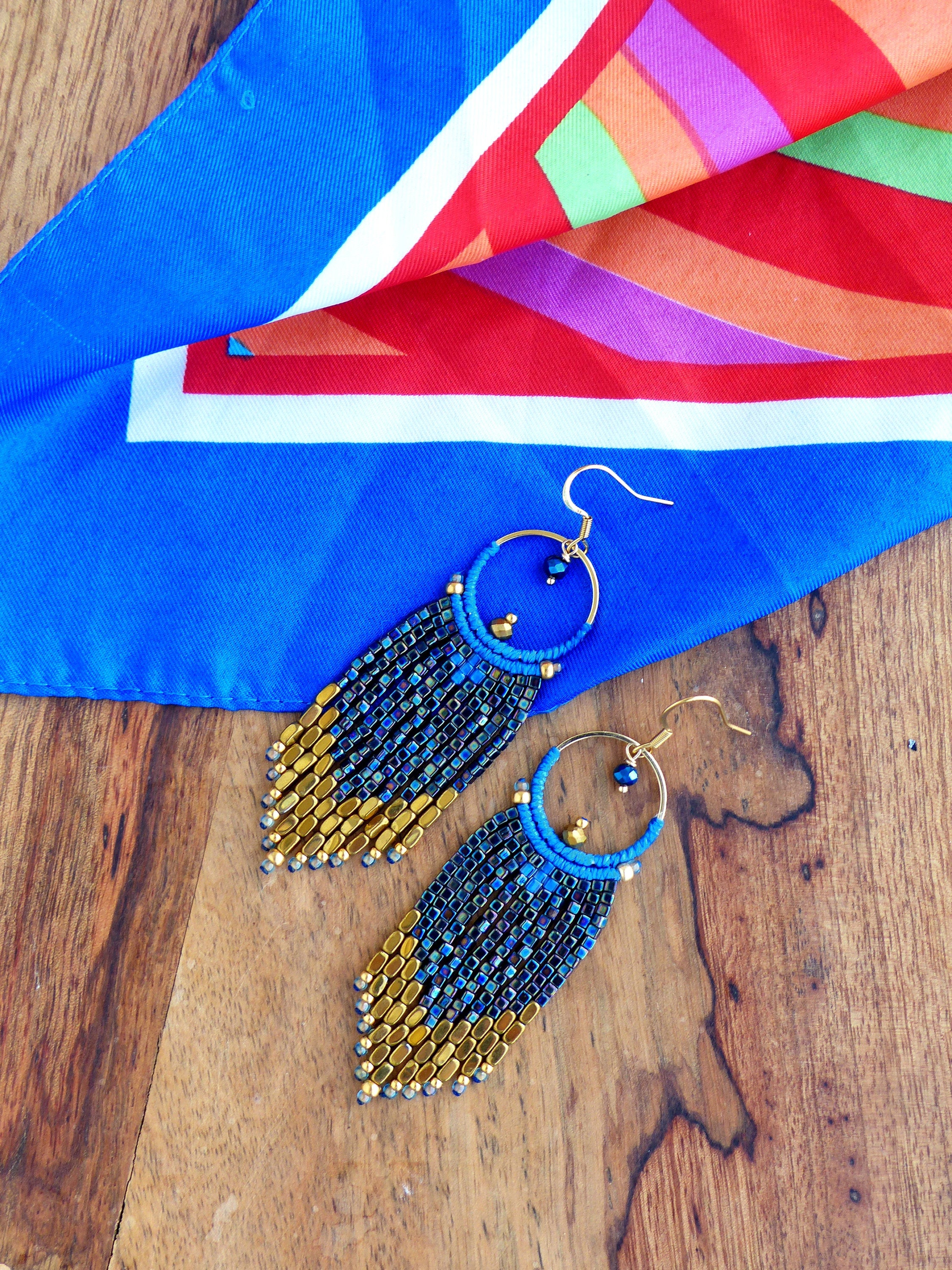 Peut inclure: Boucles d'oreilles créoles dorées avec des franges de perles bleues et dorées. Elles sont posées sur un foulard bleu et multicolore, sur une surface en bois. Les boucles d'oreilles ont un diamètre d'environ 3,8 cm.