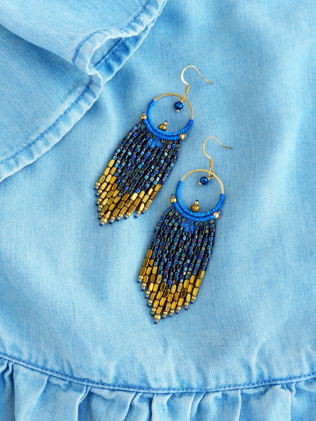 Peut inclure: Une paire de boucles d'oreilles faites à la main avec un motif de franges de perles bleues et dorées. Les boucles d'oreilles comportent des crochets dorés et un cercle bleu avec une petite perle. La frange passe de l'or au bleu foncé.
