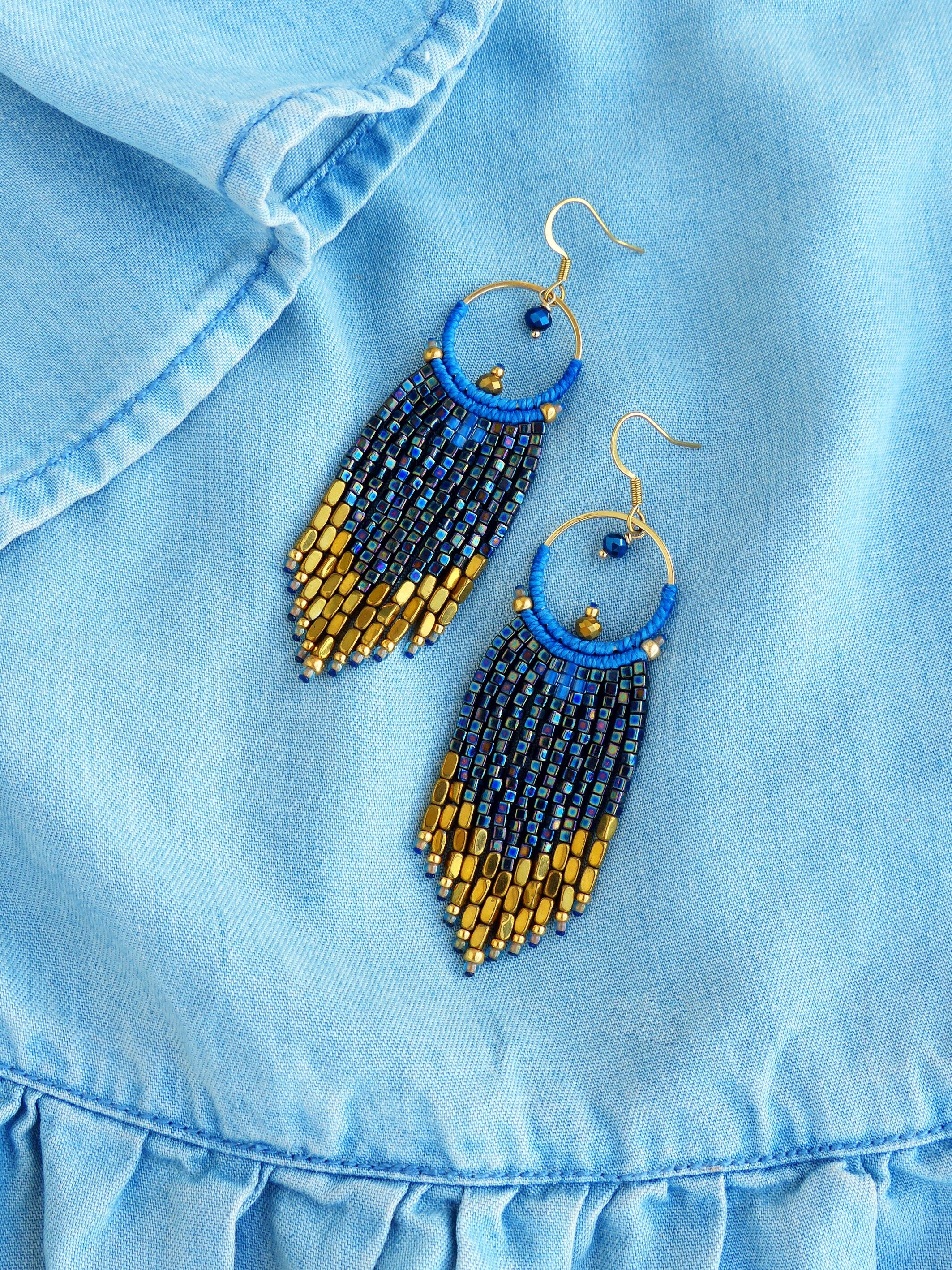 Peut inclure: Une paire de boucles d'oreilles faites à la main avec un motif de franges de perles bleues et dorées. Les boucles d'oreilles comportent des crochets dorés et un cercle bleu avec une petite perle. La frange passe de l'or au bleu foncé.