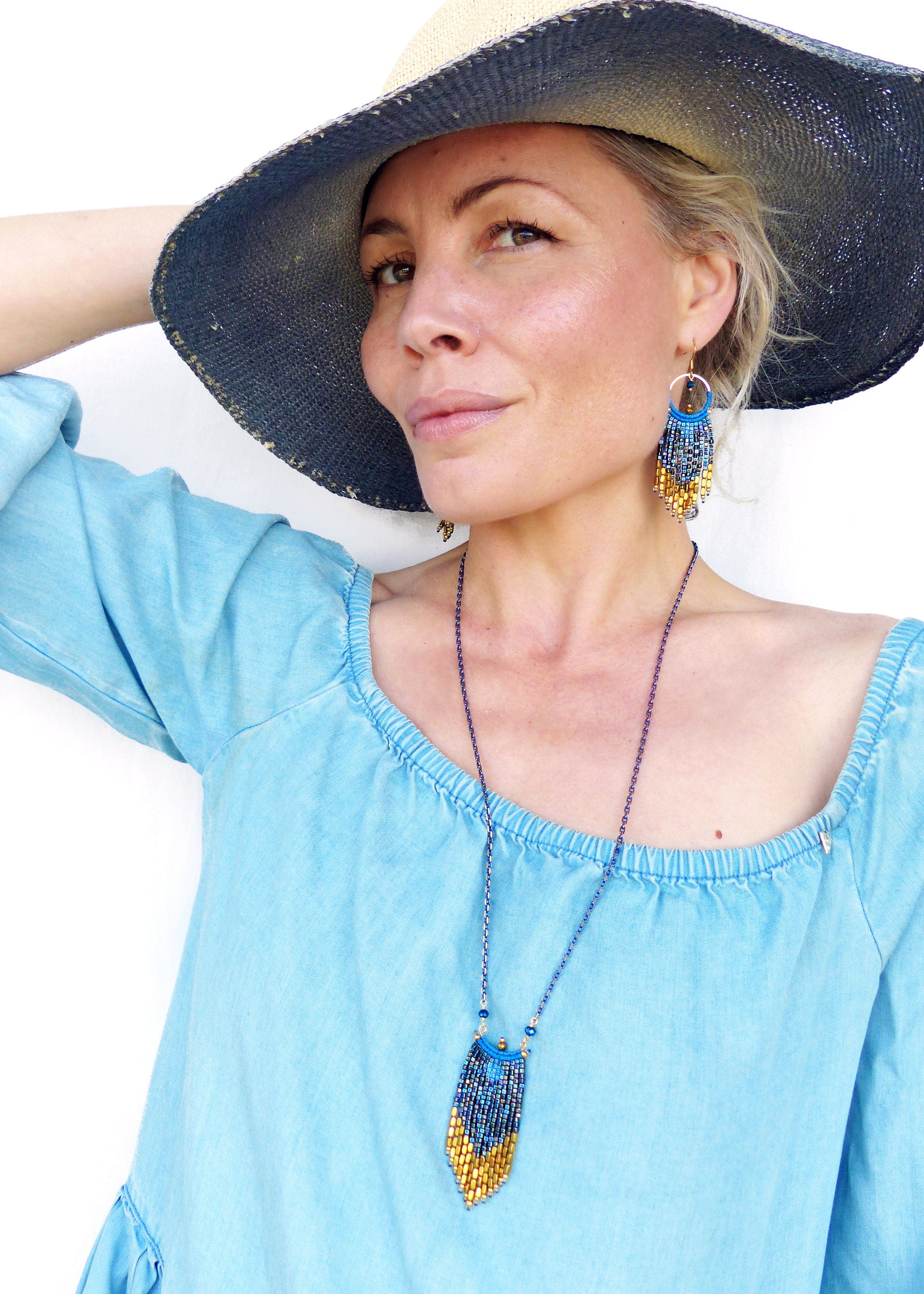 Peut inclure: Une femme porte un chapeau de paille à larges bords avec un bandeau bleu marine et un haut bleu clair à épaules dénudées. Elle porte également des boucles d'oreilles et un collier de perles avec un pendentif bleu et or.