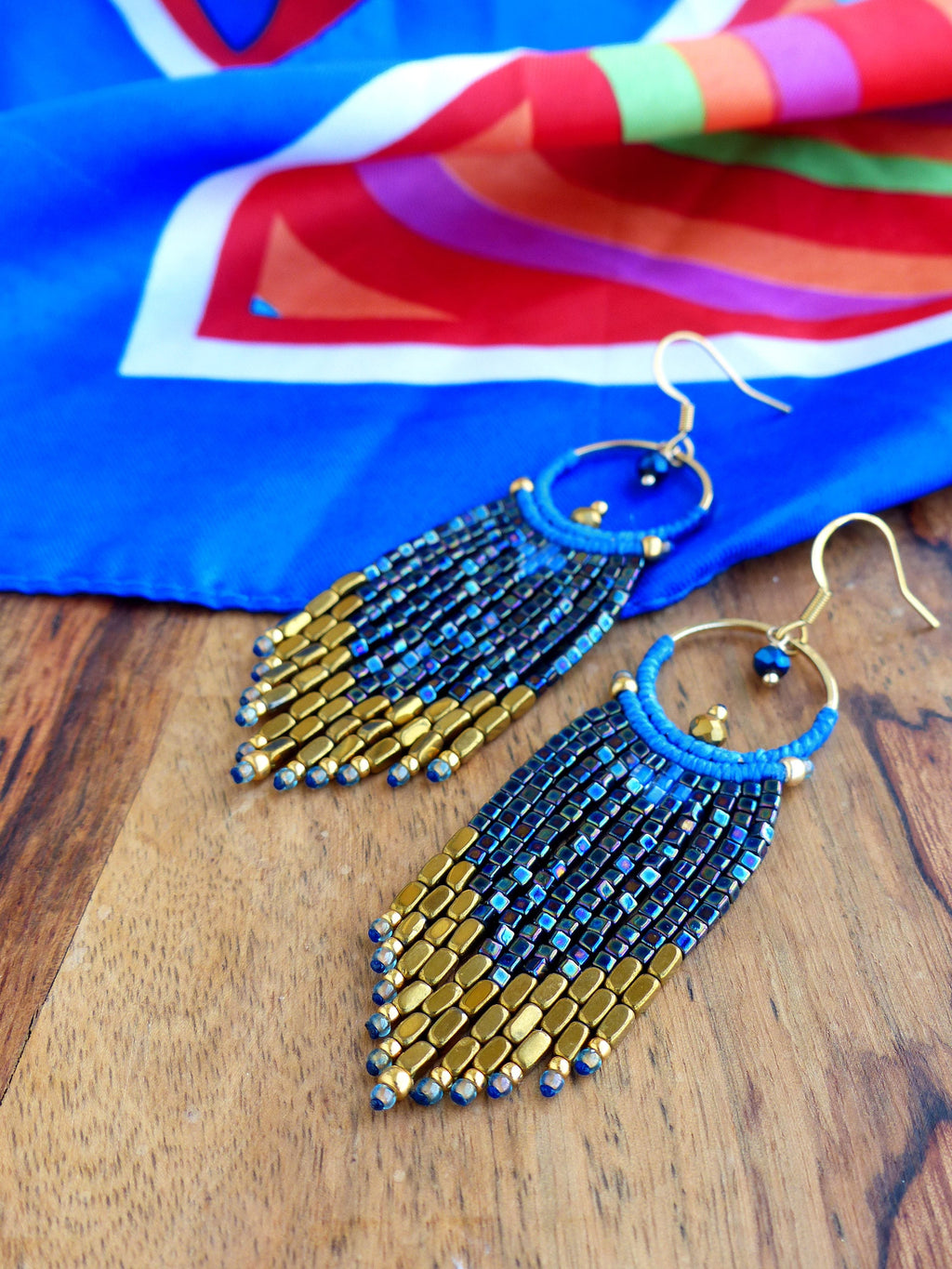 Peut inclure: Boucles d'oreilles perlées faites à la main. Elles présentent des cerceaux dorés et des détails bleus, avec une frange de petites perles bleu foncé et dorées. Elles sont présentées sur une surface en bois, avec un tissu bleu et à motifs en arrière-plan.