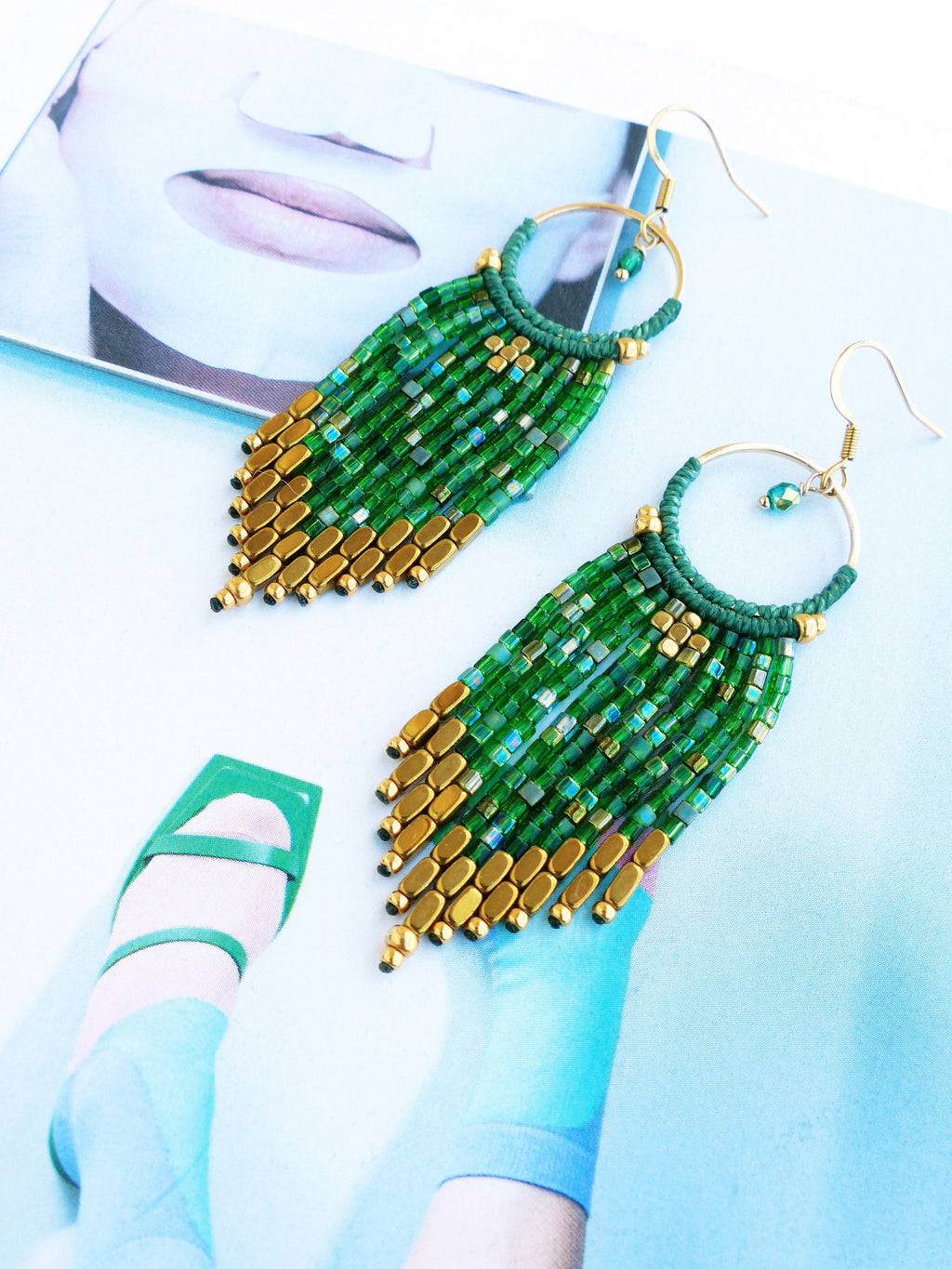 Peut inclure: Une paire de longues boucles d'oreilles en perles vertes et dorées avec un anneau en haut. Les boucles d'oreilles sont faites de petites perles carrées et de plus grandes perles rectangulaires dorées.