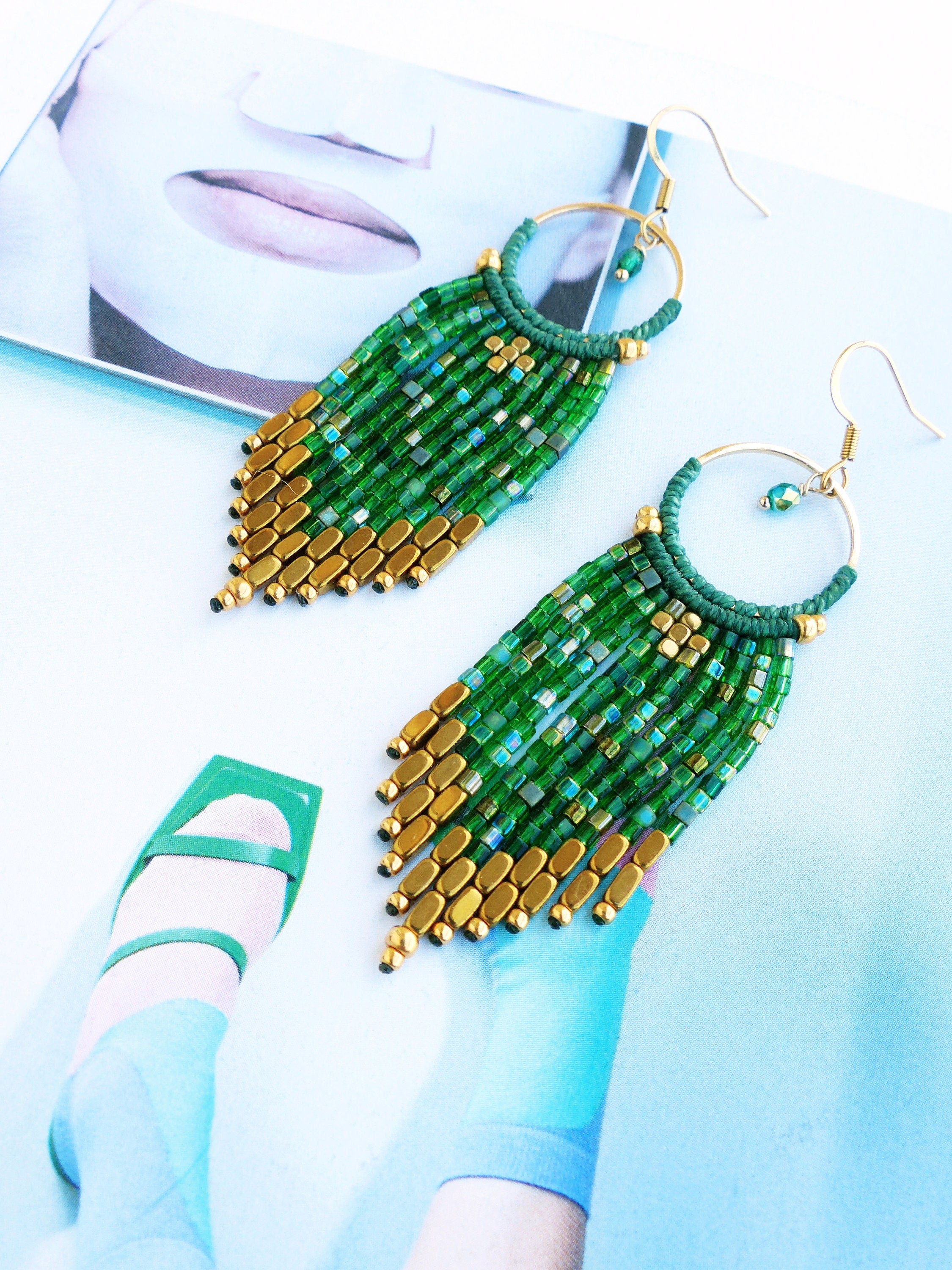 Peut inclure: Une paire de longues boucles d'oreilles en perles vertes et dorées avec un anneau en haut. Les boucles d'oreilles sont faites de petites perles carrées et de plus grandes perles rectangulaires dorées.
