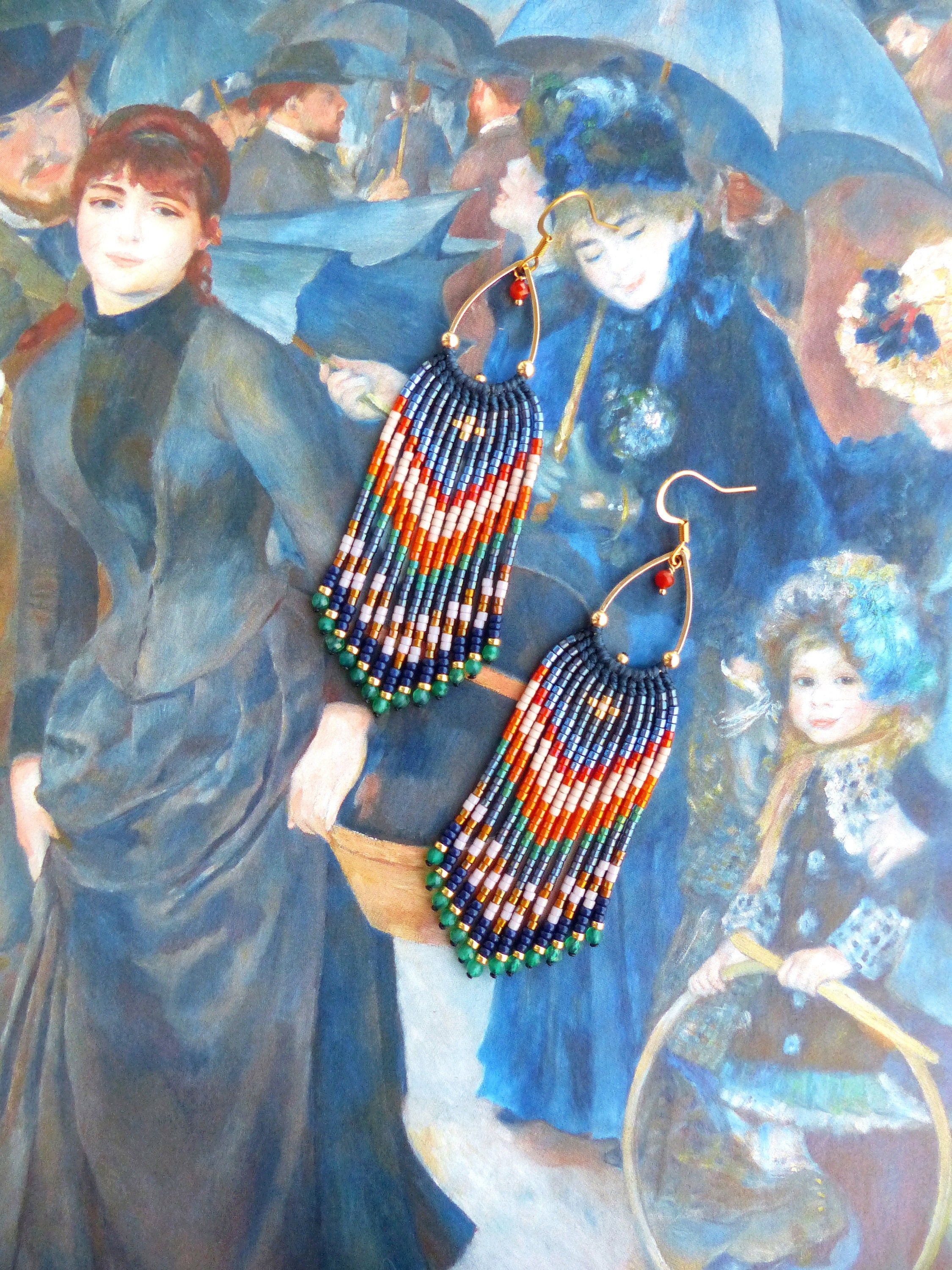 Peut inclure: Boucles d'oreilles faites à la main avec un design à franges colorées. Les boucles d'oreilles présentent un crochet doré et une cascade de perles bleues, blanches, orange, vertes et dorées. Une petite perle rouge orne le crochet. Les boucles d'oreilles sont présentées sur un fond de peinture.