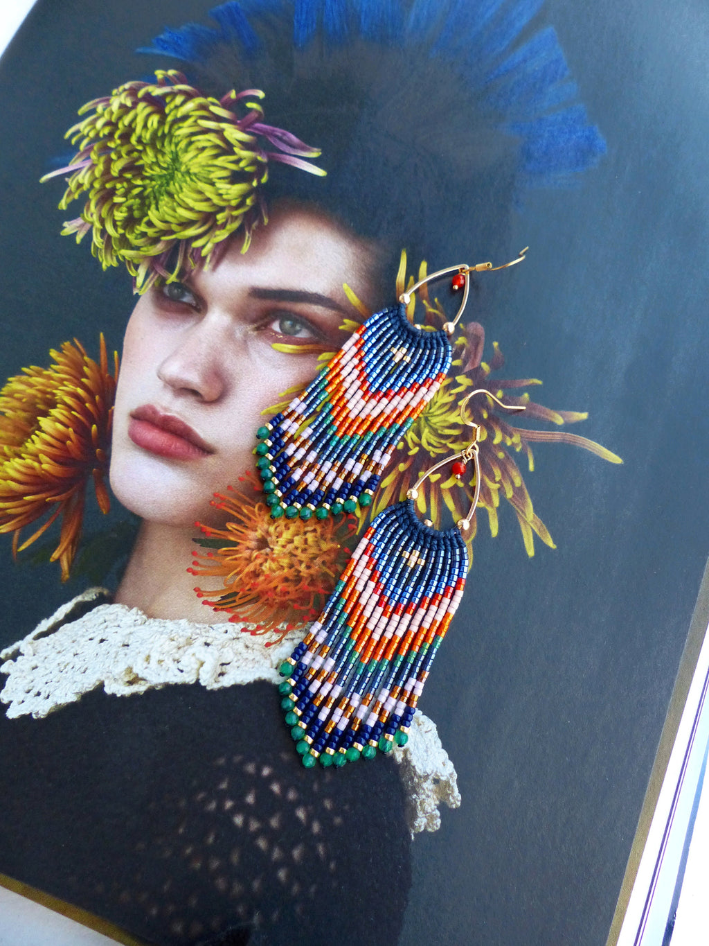 Peut inclure: Une paire de boucles d'oreilles faites à la main avec un motif de franges. Les boucles d'oreilles ont des cerceaux dorés et sont ornées d'un motif de perles bleues, oranges, blanches et vertes. Les boucles d'oreilles sont présentées sur un magazine.