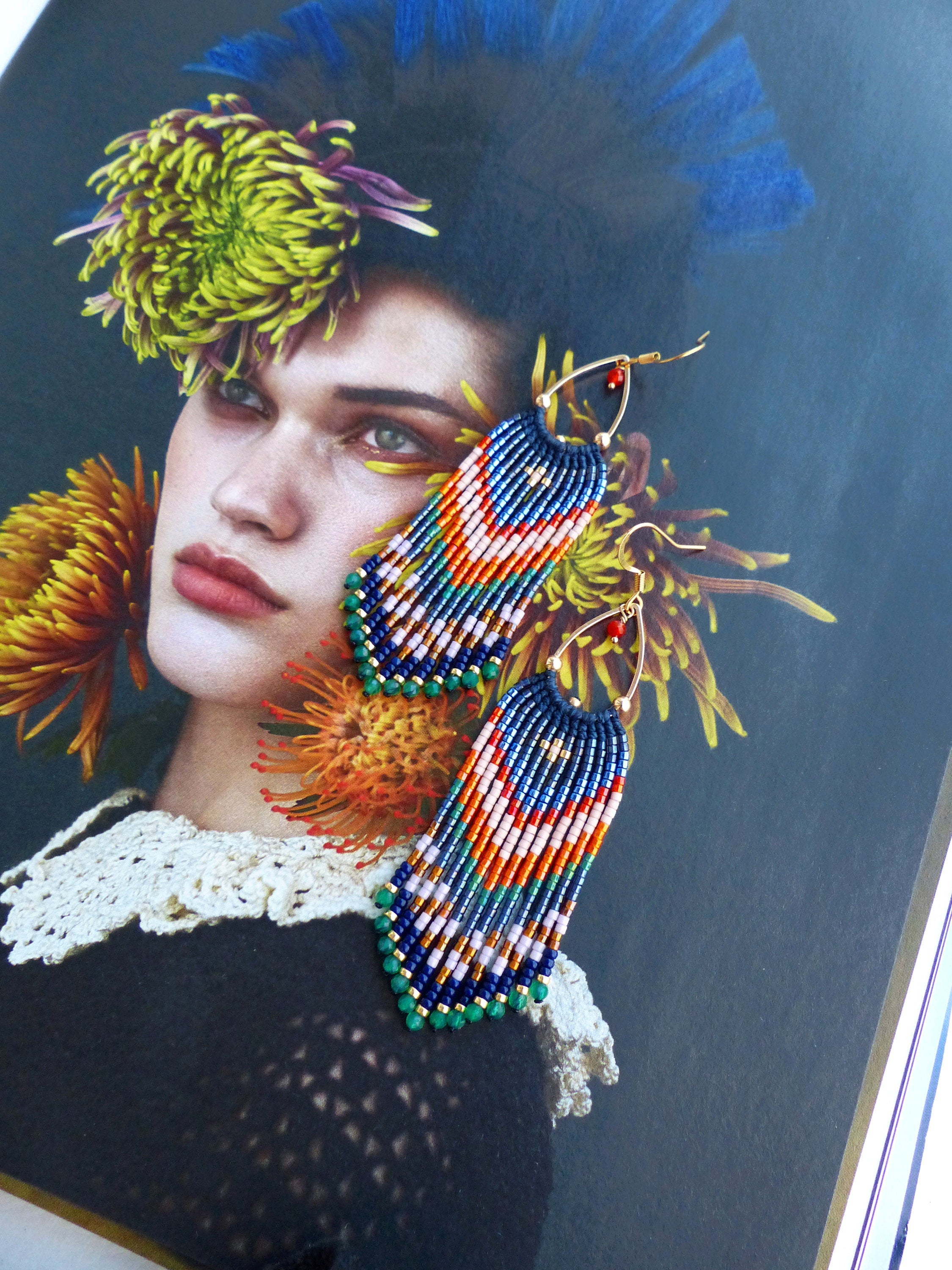 Peut inclure: Une paire de boucles d'oreilles faites à la main avec un motif de franges. Les boucles d'oreilles ont des cerceaux dorés et sont ornées d'un motif de perles bleues, oranges, blanches et vertes. Les boucles d'oreilles sont présentées sur un magazine.