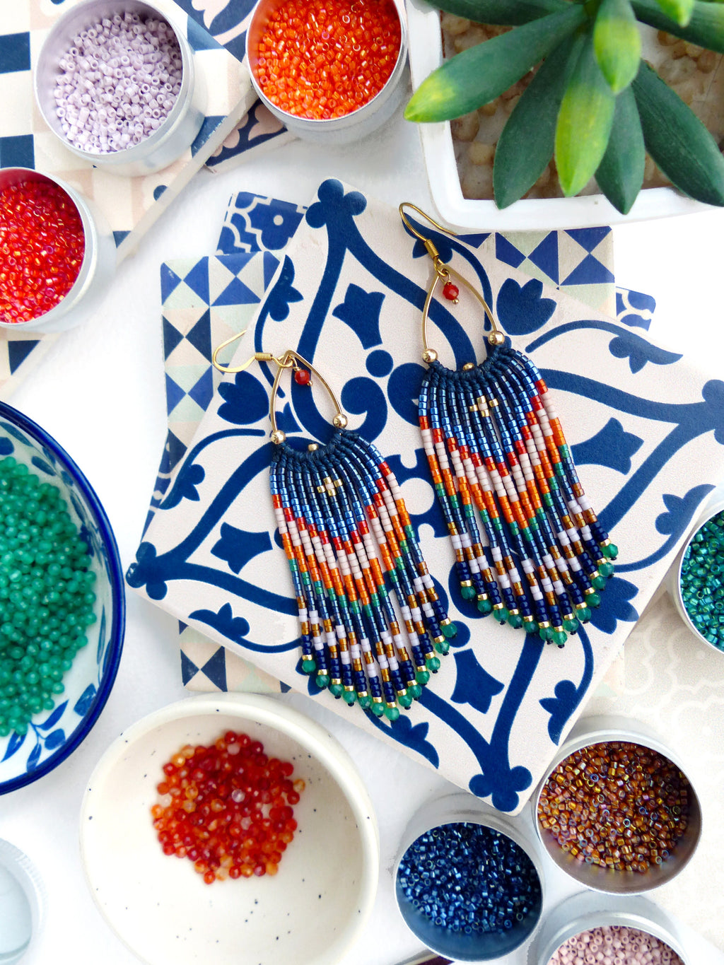 Peut inclure: Boucles d'oreilles artisanales en perles avec une partie supérieure bleu marine et des franges colorées. Les boucles d'oreilles sont présentées sur un carreau à motifs bleus et blancs. Divers petits récipients de perles rouges, oranges, vertes et violettes entourent les boucles d'oreilles.