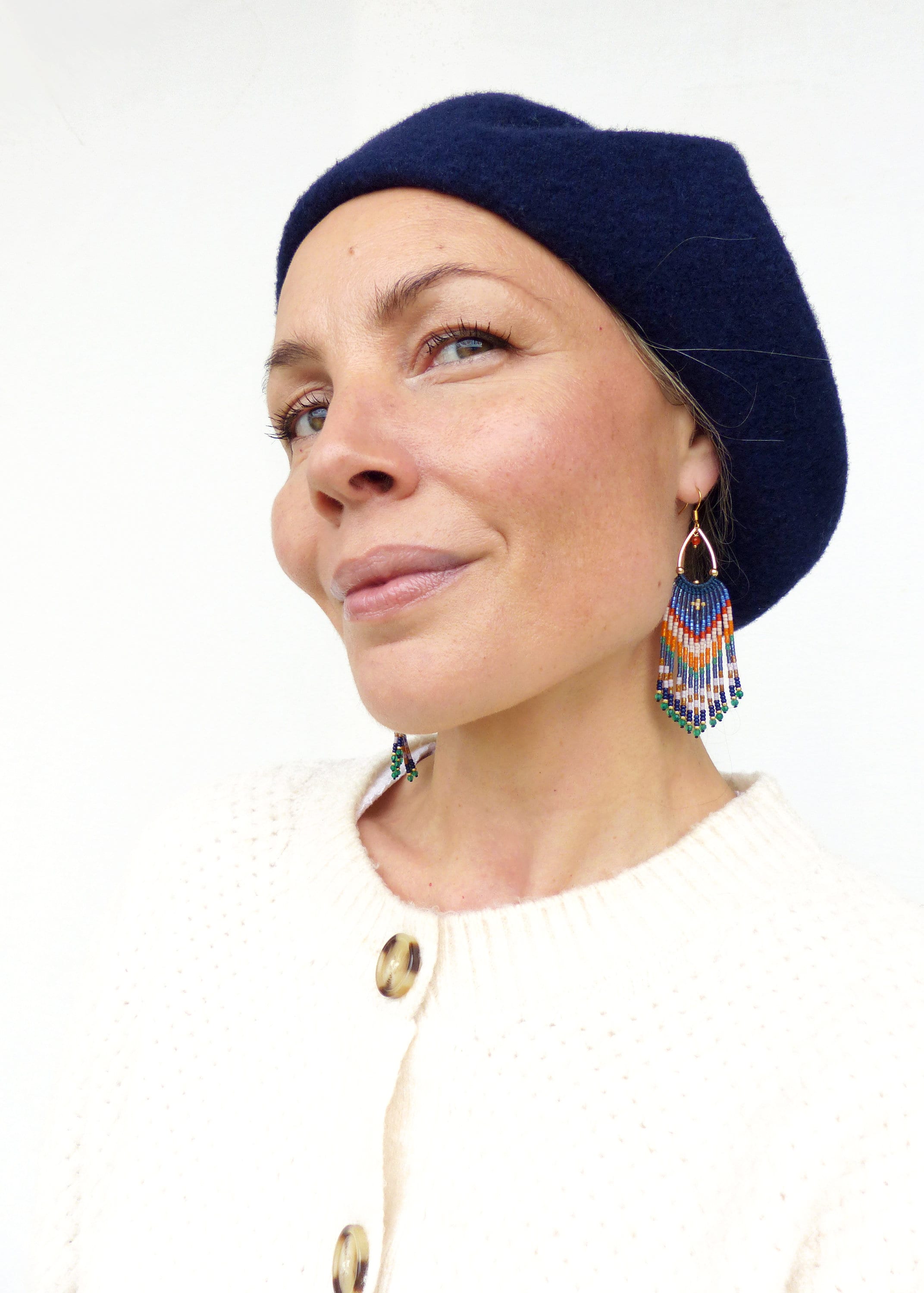 Peut inclure: Béret bleu marine et boucles d'oreilles en perles. Les boucles d'oreilles présentent un cerceau doré avec une frange de perles colorées dans les tons de bleu, orange, blanc et vert. La personne porte un cardigan crème.