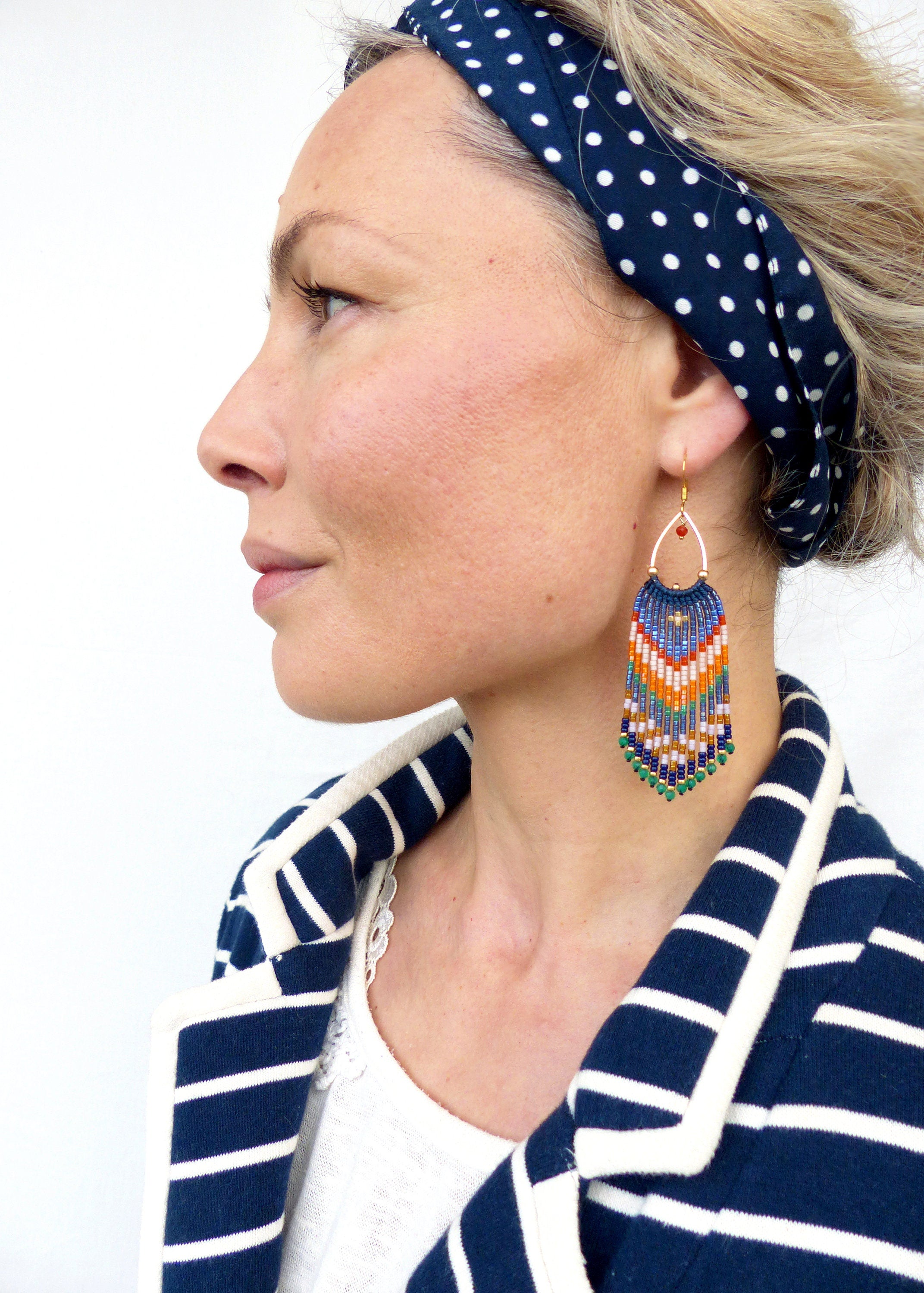 Peut inclure: Boucles d'oreilles à franges perlées bleues, oranges, vertes et blanches. Elles sont suspendues à des cerceaux dorés. La personne porte une veste rayée bleu marine et blanche et un bandeau à pois bleu marine.