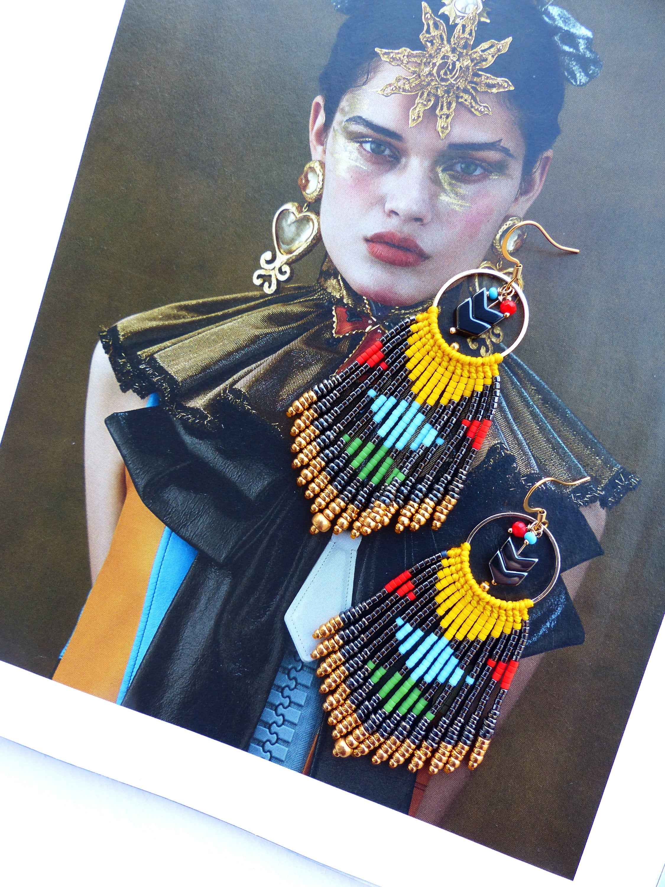 Peut inclure: Une paire de longues boucles d'oreilles en perles avec un motif géométrique. Les boucles d'oreilles sont faites de perles noires, jaunes, rouges, vertes et bleues. Les boucles d'oreilles sont suspendues à des cercles dorés.
