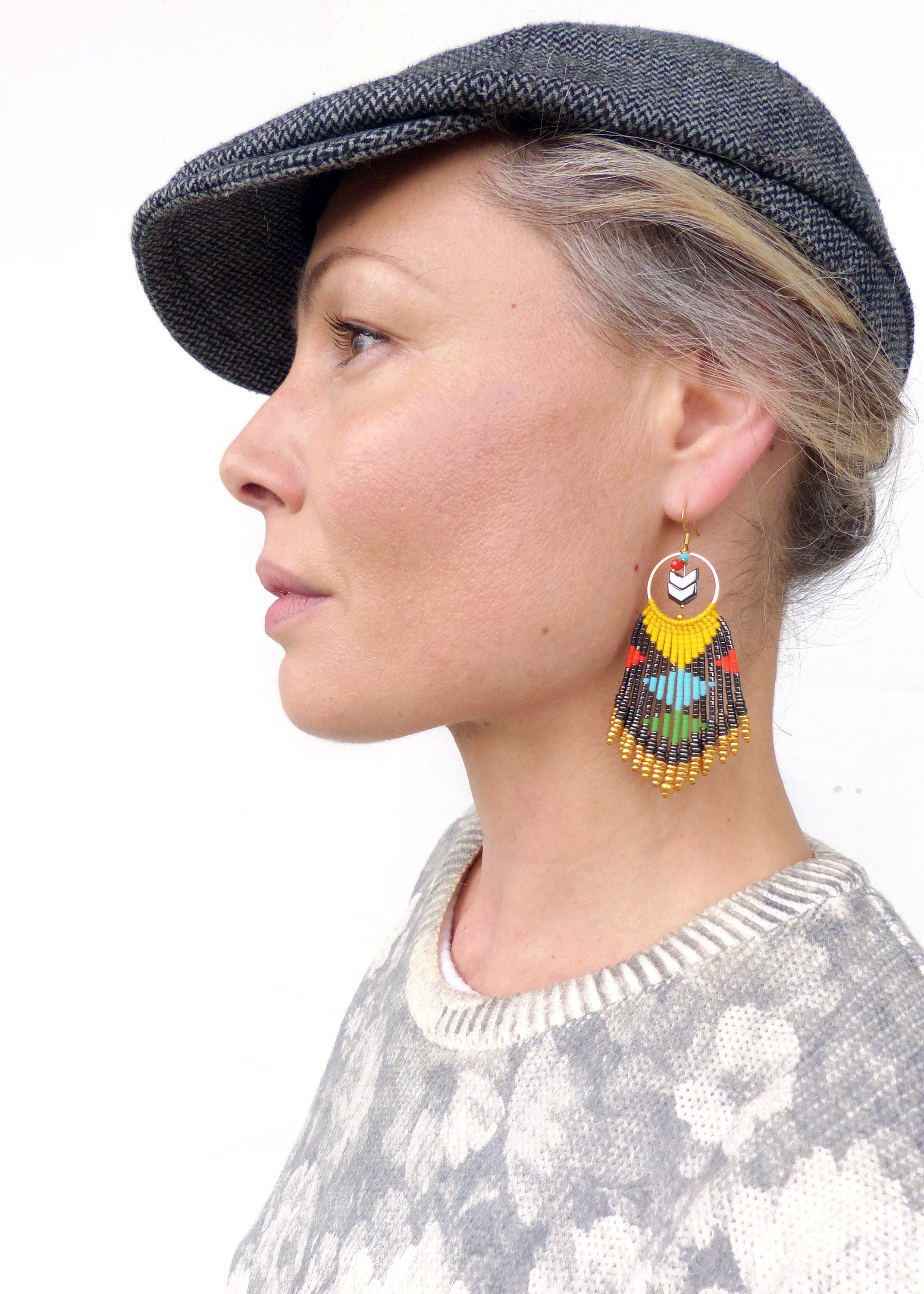 Peut inclure: Une femme portant un pull à motifs gris et noir et une casquette de newsboy en tweed gris. Elle porte une paire de longues boucles d'oreilles en perles colorées avec un anneau en or et un motif de flèche blanche.