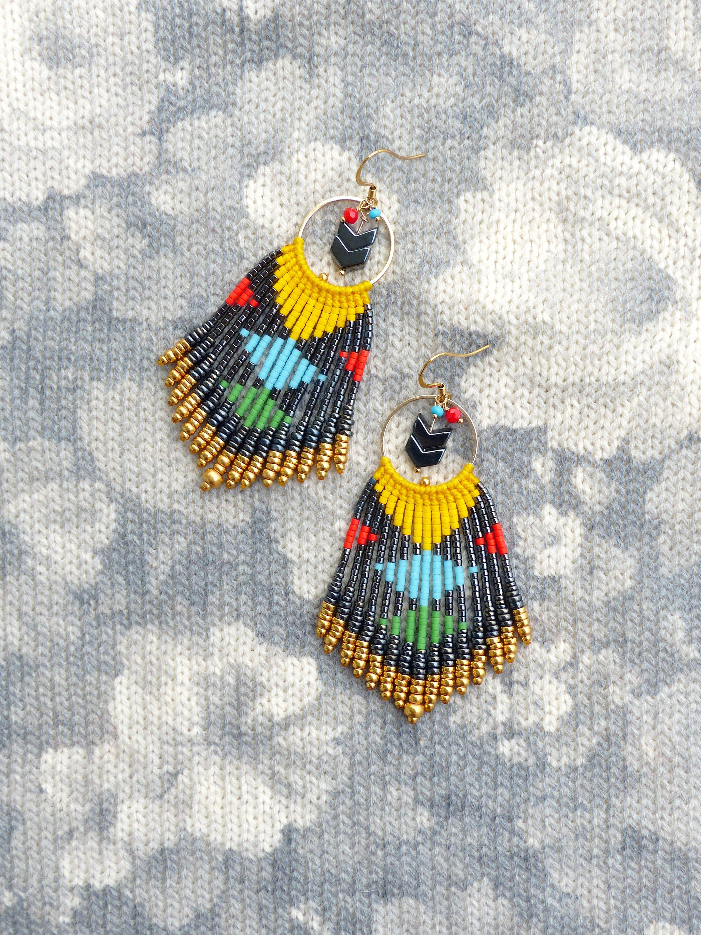 Peut inclure: Une paire de longues boucles d'oreilles en perles avec un anneau en or et un pendentif en forme de flèche noire. Les boucles d'oreilles sont faites de perles noires, dorées, jaunes, rouges, bleues et vertes.