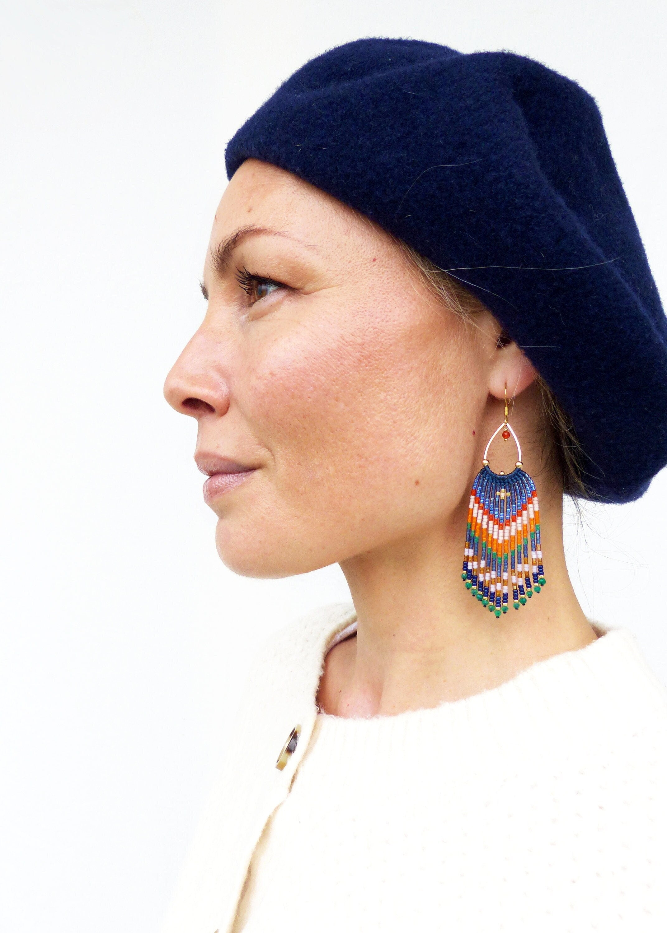 Peut inclure: Une femme porte un béret bleu marine et des boucles d'oreilles en perles colorées. Les boucles d'oreilles ont un cerceau doré et présentent un motif géométrique bleu, orange, blanc et vert, avec des franges perlées.
