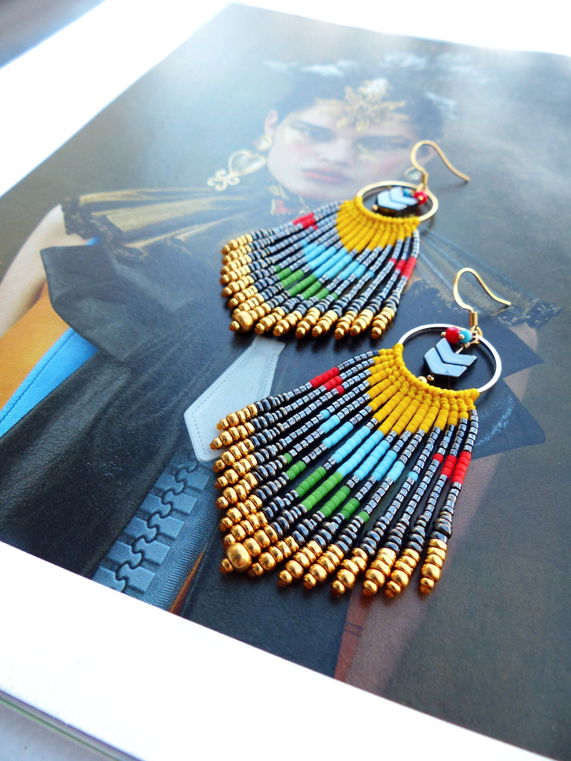 Peut inclure: Une paire de longues boucles d'oreilles en perles colorées avec des accents dorés. Les boucles d'oreilles présentent une frange de perles dans des tons de bleu, vert, jaune, rouge et noir, avec des perles dorées entremêlées. Les boucles d'oreilles sont suspendues à des anneaux dorés avec un motif de flèche noire.