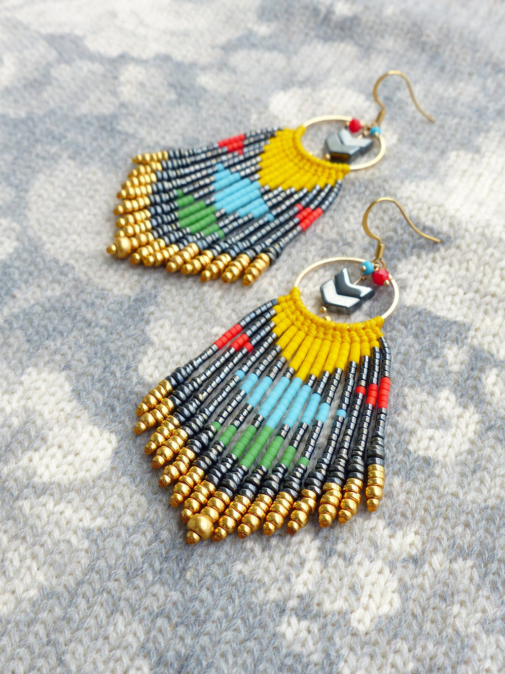 Peut inclure: Une paire de longues boucles d'oreilles en perles colorées avec des accents dorés. Les boucles d'oreilles présentent un schéma de couleurs jaune, bleu, vert, rouge et noir. Les boucles d'oreilles sont suspendues à des cercles dorés avec un motif de flèche noire.