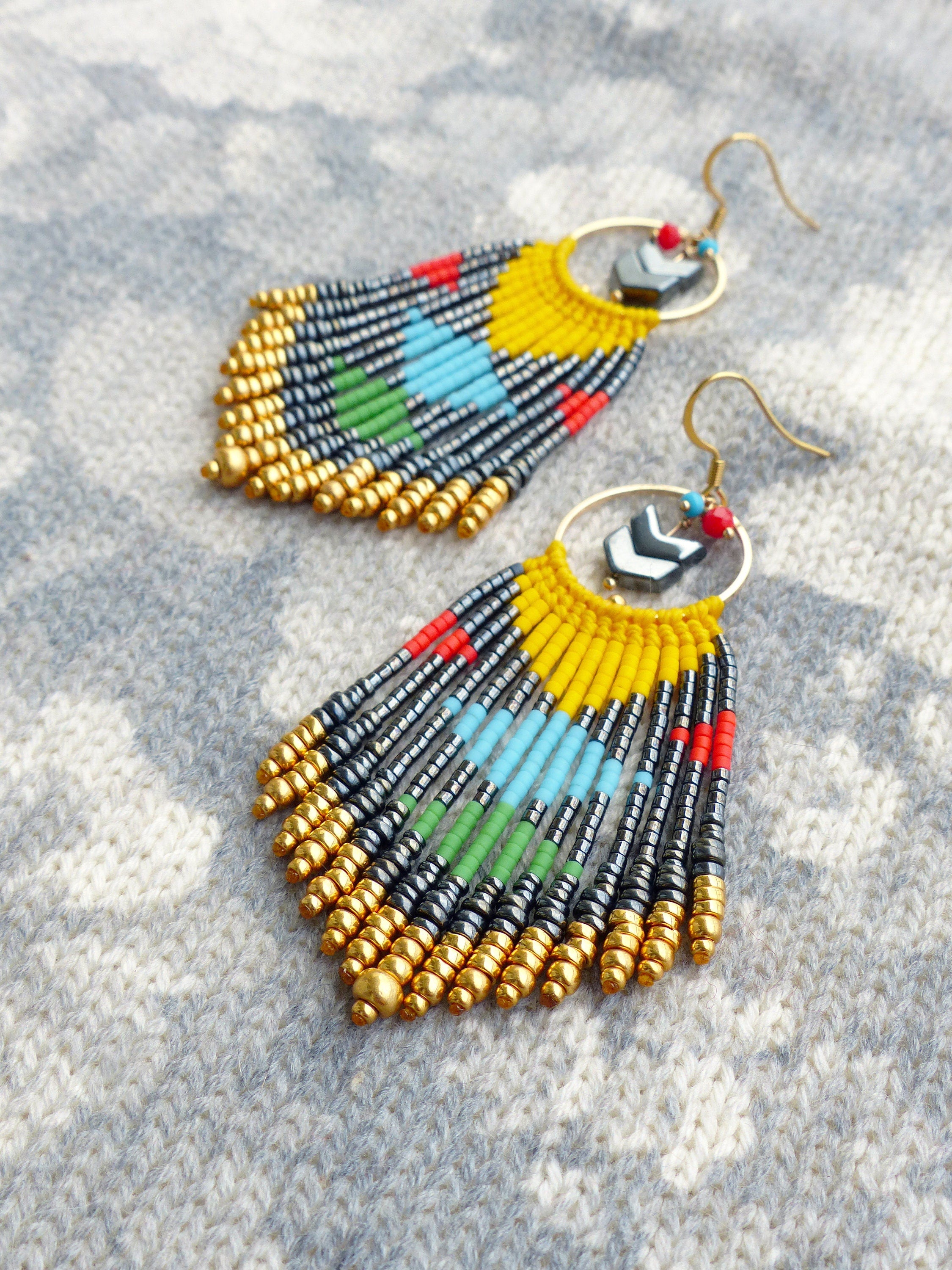 Peut inclure: Une paire de longues boucles d'oreilles en perles colorées avec des accents dorés. Les boucles d'oreilles présentent un schéma de couleurs jaune, bleu, vert, rouge et noir. Les boucles d'oreilles sont suspendues à des cercles dorés avec un motif de flèche noire.