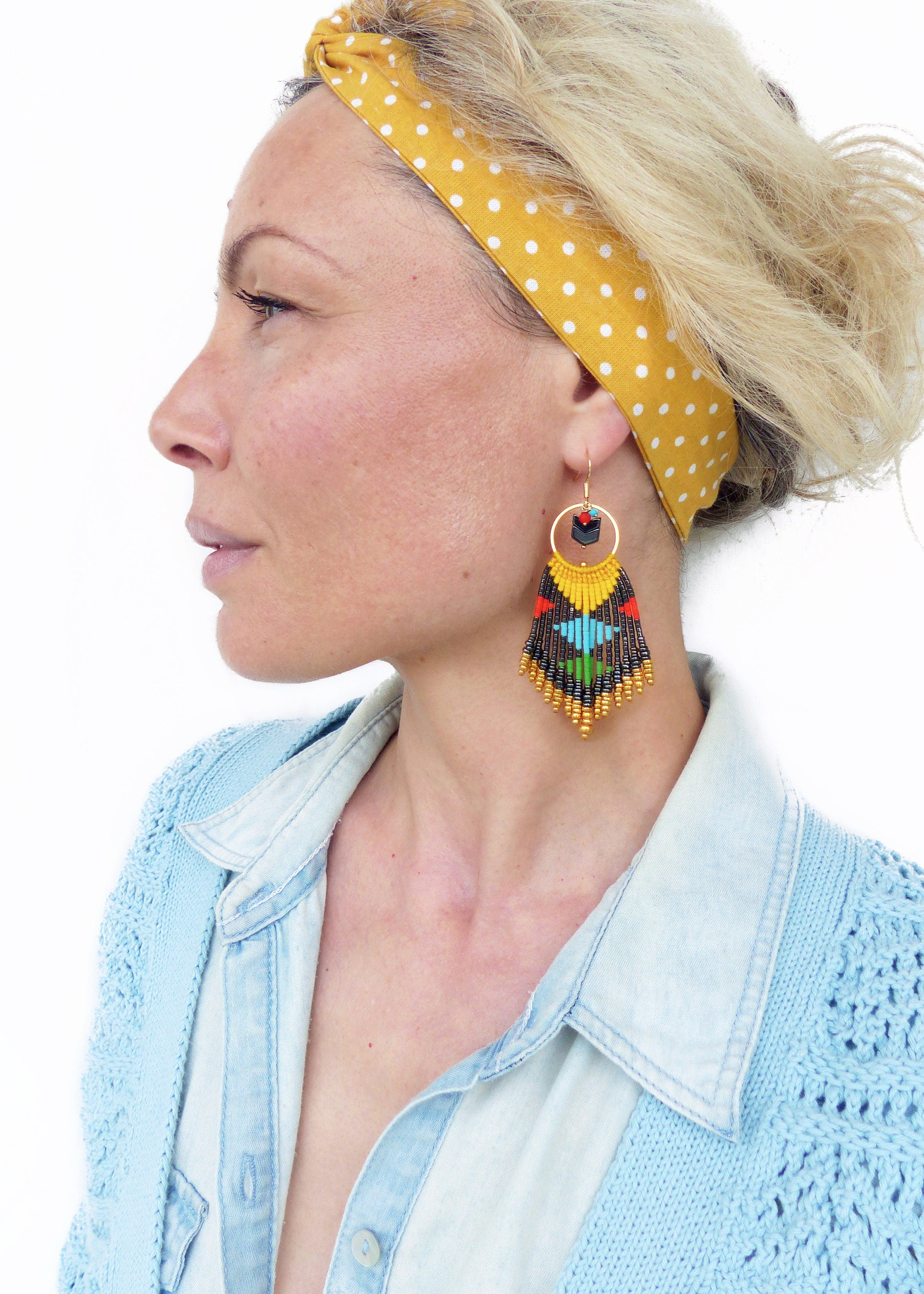 Peut inclure: Une femme portant un bandeau jaune à pois blancs et un cardigan bleu. Elle porte de longues boucles d'oreilles en perles colorées avec un motif géométrique noir et or.