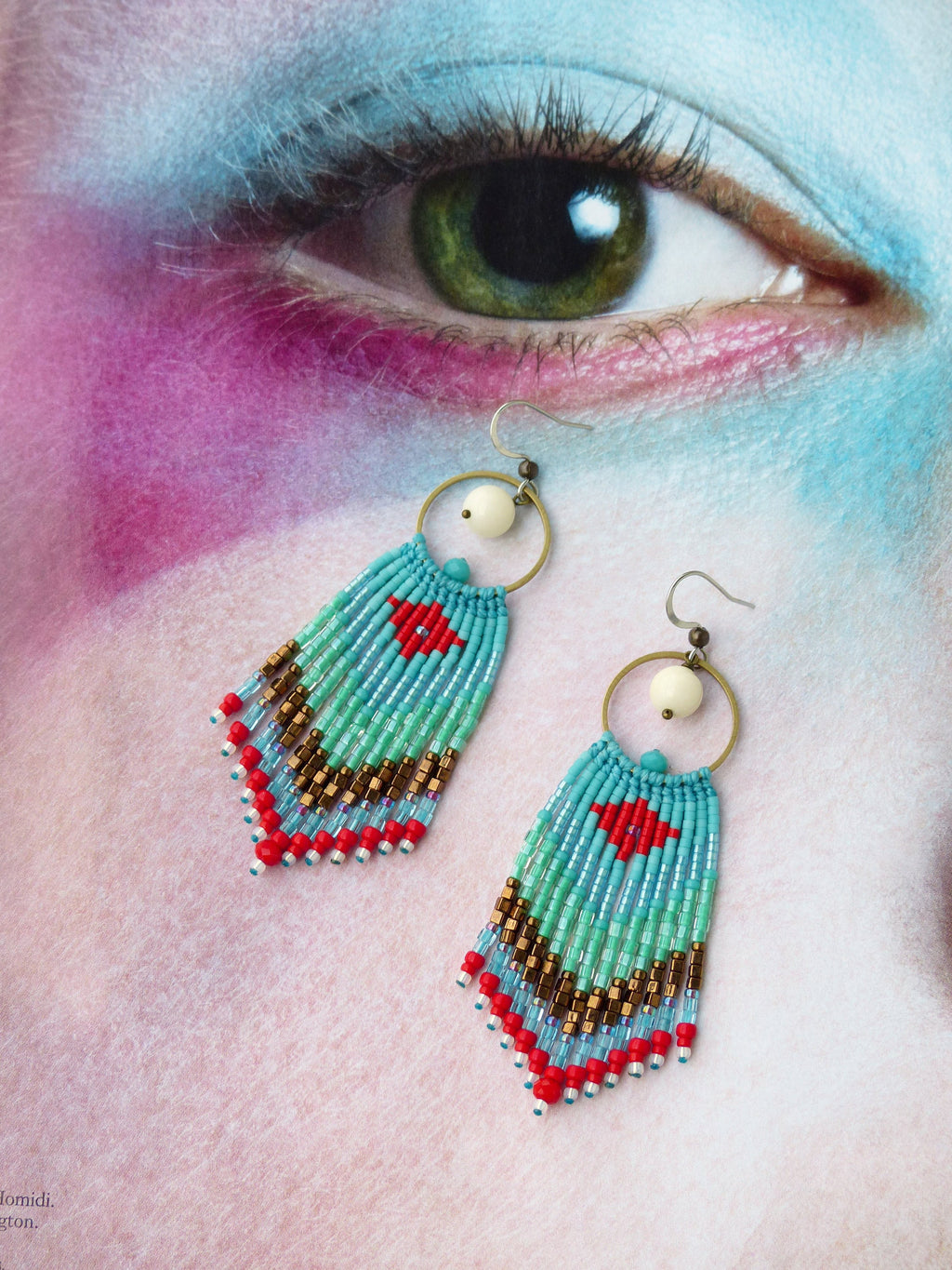 Peut inclure: Une paire de boucles d'oreilles en perles turquoise, rouge et or avec une perle blanche et un anneau doré en haut. Les boucles d'oreilles pendent sur un fond blanc.