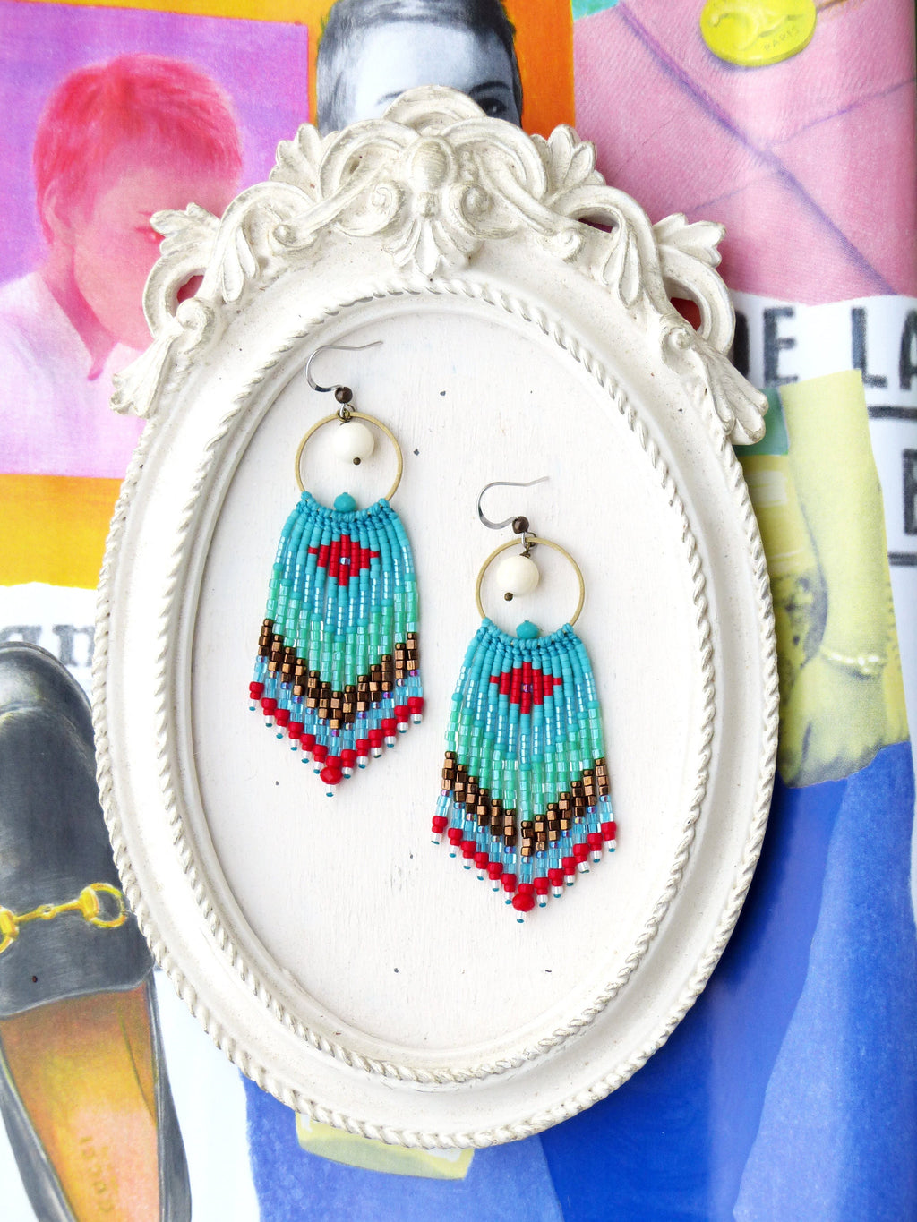 Peut inclure: Une paire de boucles d'oreilles en perles turquoise, rouge et blanche avec des cercles dorés et des perles blanches suspendues aux cercles. Les boucles d'oreilles sont présentées sur un cadre ovale blanc.