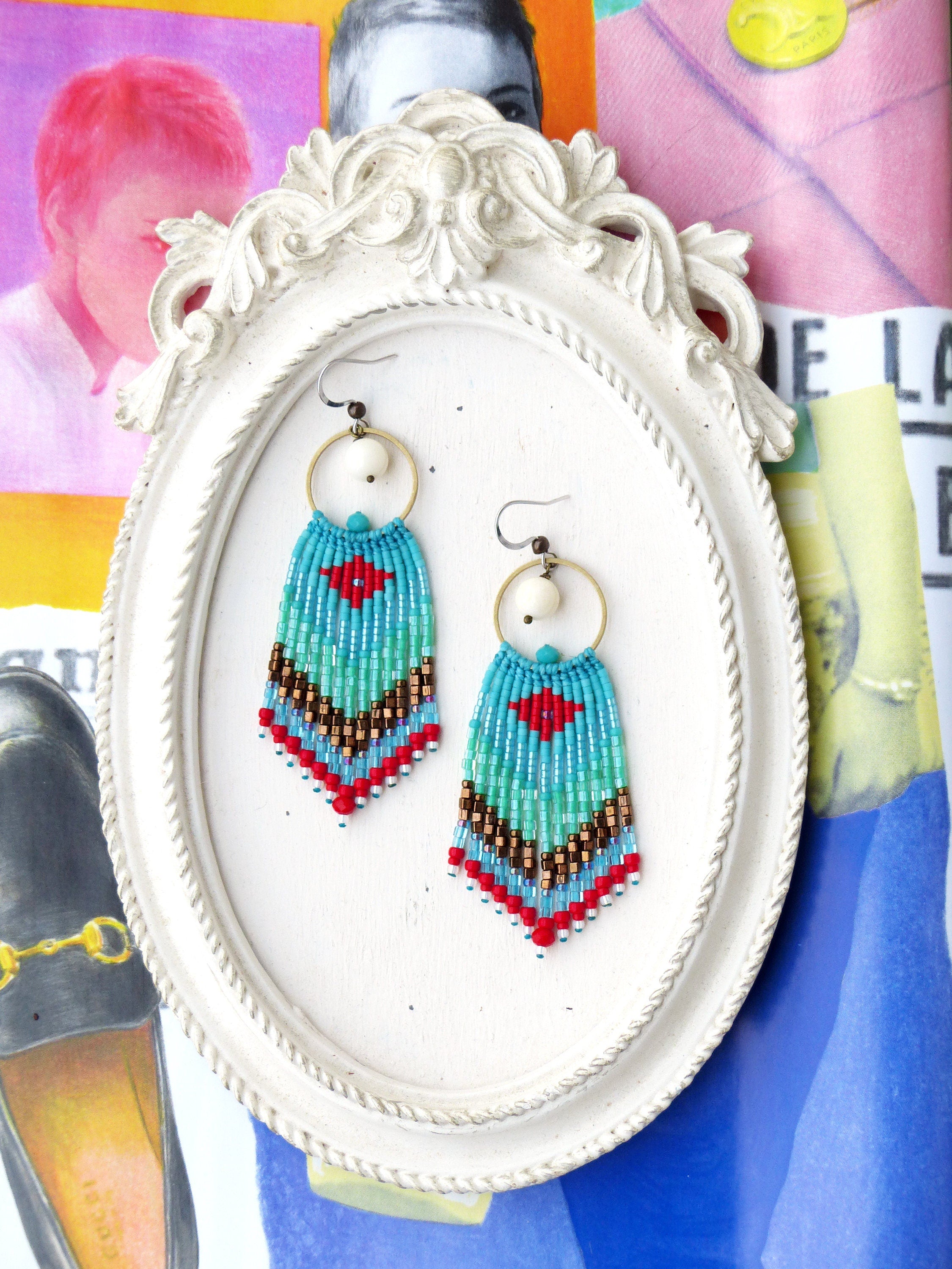Peut inclure: Une paire de boucles d'oreilles en perles turquoise, rouge et blanche avec des cercles dorés et des perles blanches suspendues aux cercles. Les boucles d'oreilles sont présentées sur un cadre ovale blanc.