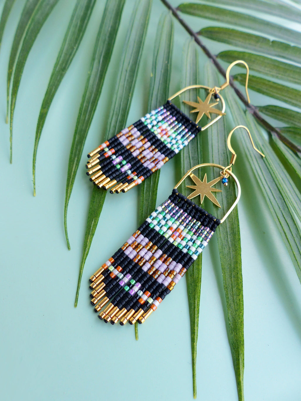 Peut inclure: Une paire de longues boucles d'oreilles en perles avec un charme d'étoile dorée. Les boucles d'oreilles sont faites de perles noires, blanches, orange, violettes et vertes. Les boucles d'oreilles sont suspendues sur un fond bleu clair.