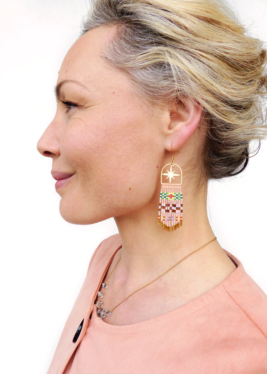 Peut inclure: Une femme portant un chemisier couleur pêche avec un collier en chaîne dorée et des accents de cristal. Elle porte de longues boucles d'oreilles en perles avec un motif d'étoile dorée.
