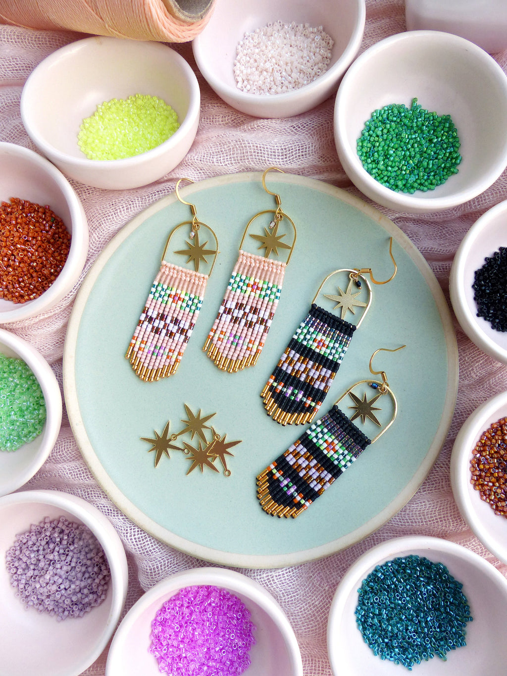 Peut inclure: Une paire de boucles d'oreilles en perles faites à la main avec des accents d'étoiles dorées. Les boucles d'oreilles sont faites avec des perles de rocaille colorées et présentent un design en frange. Les boucles d'oreilles sont présentées sur une assiette avec de petits bols de perles de différentes couleurs.