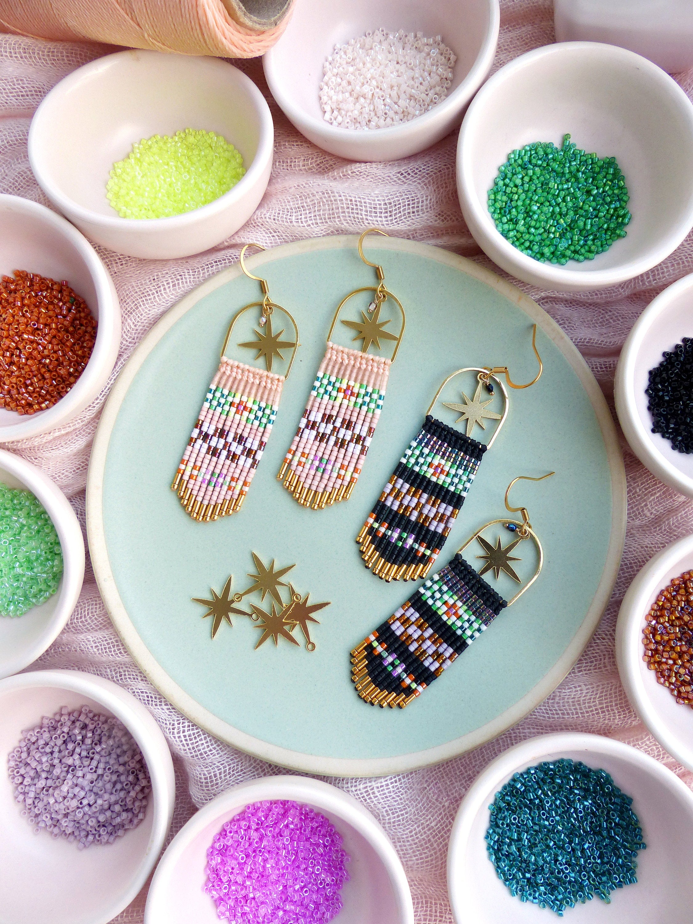 Peut inclure: Une paire de boucles d'oreilles en perles faites à la main avec des accents d'étoiles dorées. Les boucles d'oreilles sont faites avec des perles de rocaille colorées et présentent un design en frange. Les boucles d'oreilles sont présentées sur une assiette avec de petits bols de perles de différentes couleurs.