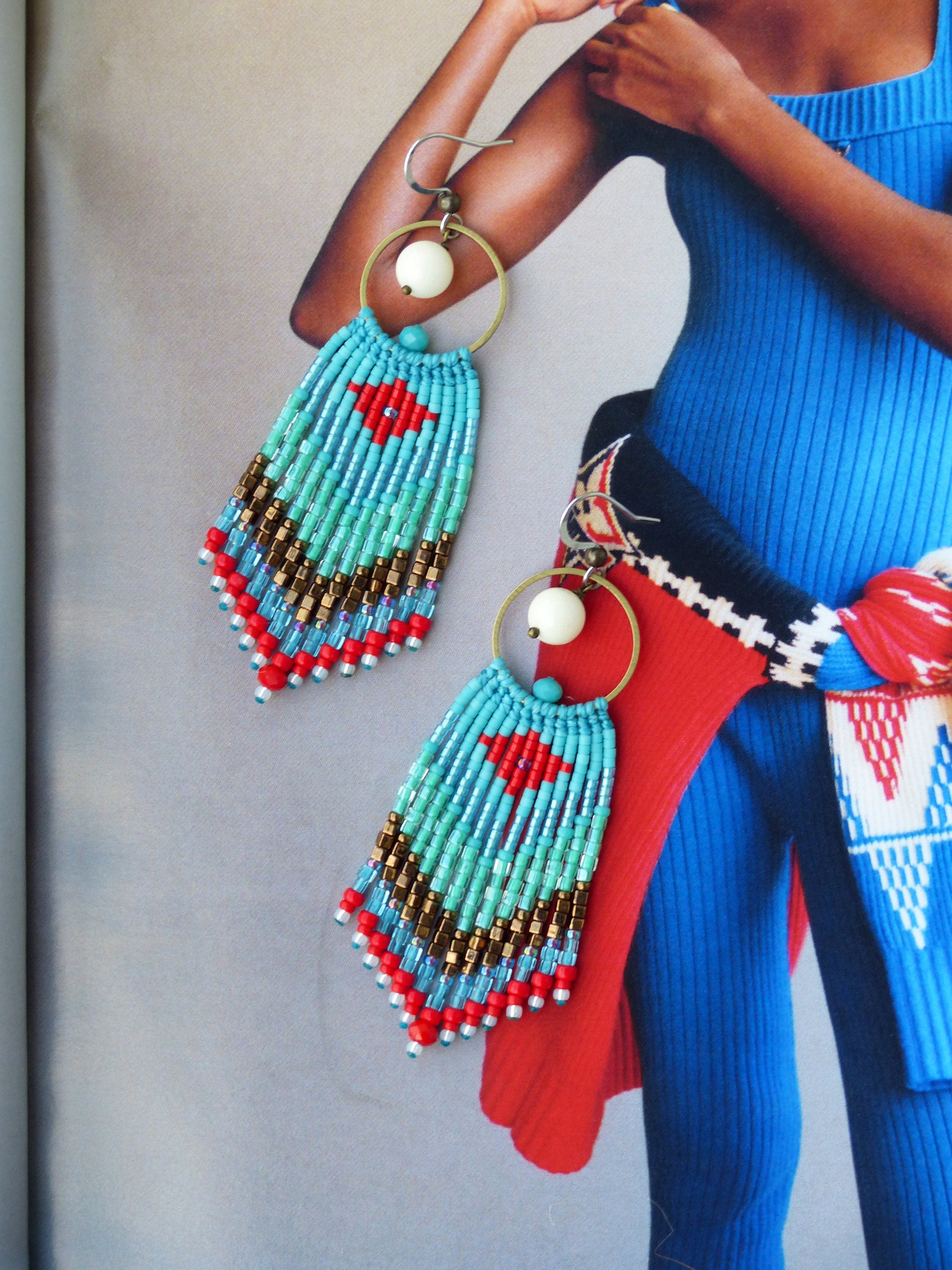 Peut inclure: Une paire de boucles d'oreilles en perles turquoise et rouge avec une perle blanche au sommet de chaque boucle d'oreille. Les boucles d'oreilles sont suspendues à un anneau en or.