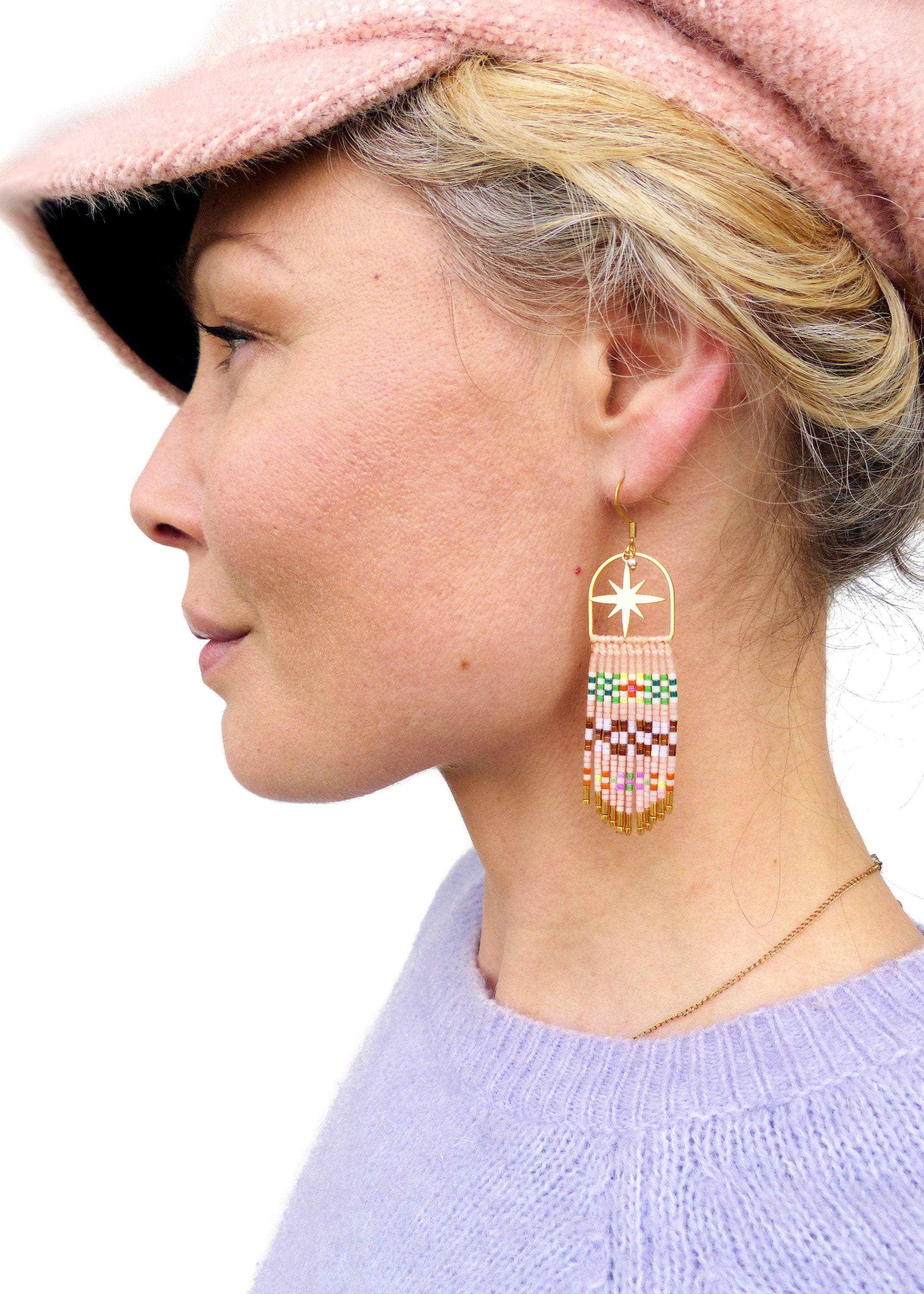 Peut inclure: Une paire de boucles d'oreilles en or avec une découpe en forme d'étoile et une frange de perles colorées. Les boucles d'oreilles pendent de l'oreille d'une personne portant un chapeau rose et un pull violet.