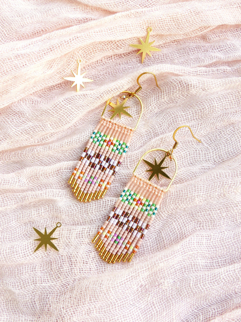 Peut inclure: Une paire de longues boucles d'oreilles en perles avec des breloques en forme d'étoile dorées. Les boucles d'oreilles sont faites de petites perles colorées et ont un design en forme de frange. Les boucles d'oreilles sont présentées sur un fond en tissu rose clair.