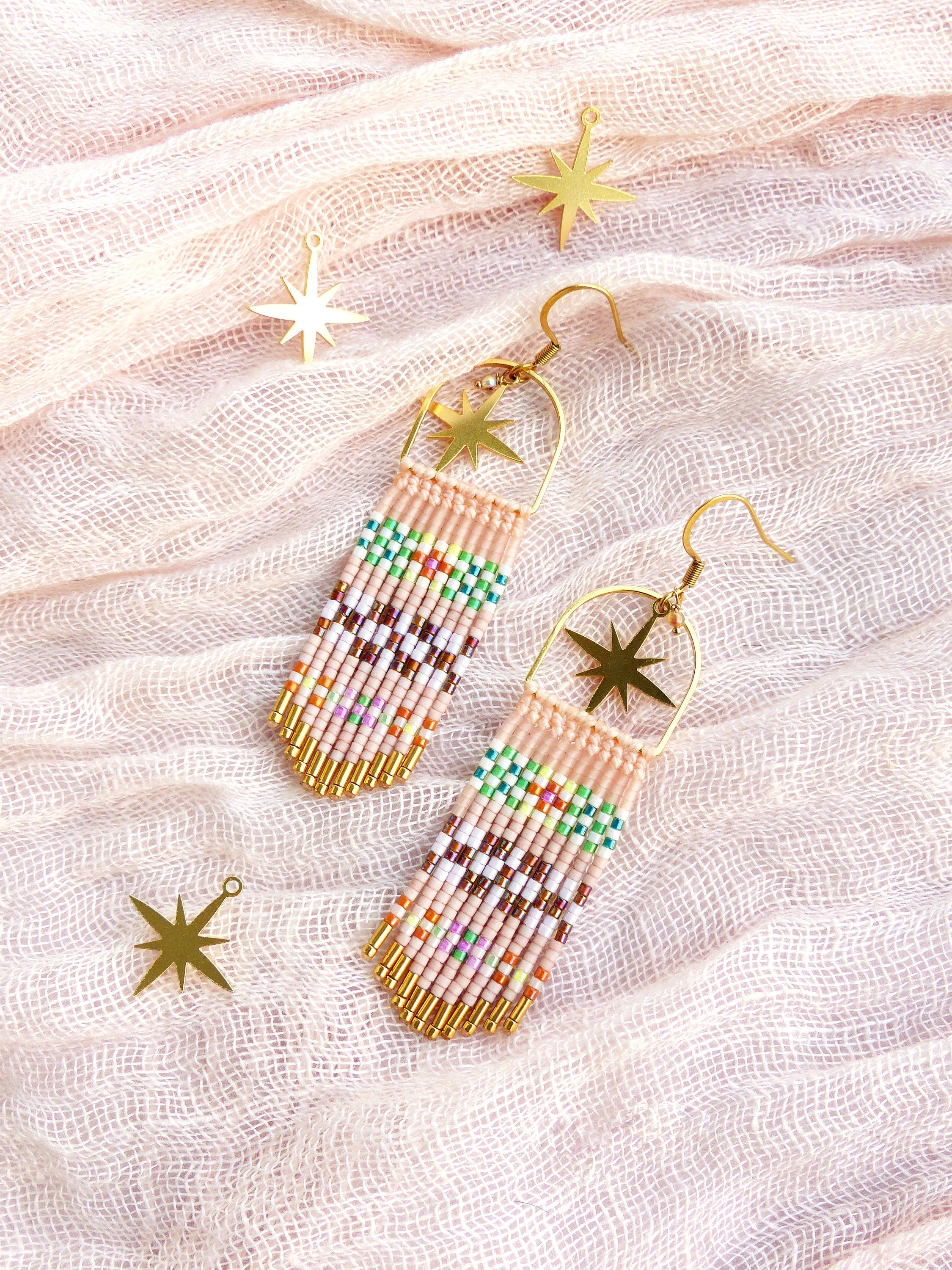 Peut inclure: Une paire de longues boucles d'oreilles en perles avec des breloques en forme d'étoile dorées. Les boucles d'oreilles sont faites de petites perles colorées et ont un design en forme de frange. Les boucles d'oreilles sont présentées sur un fond en tissu rose clair.