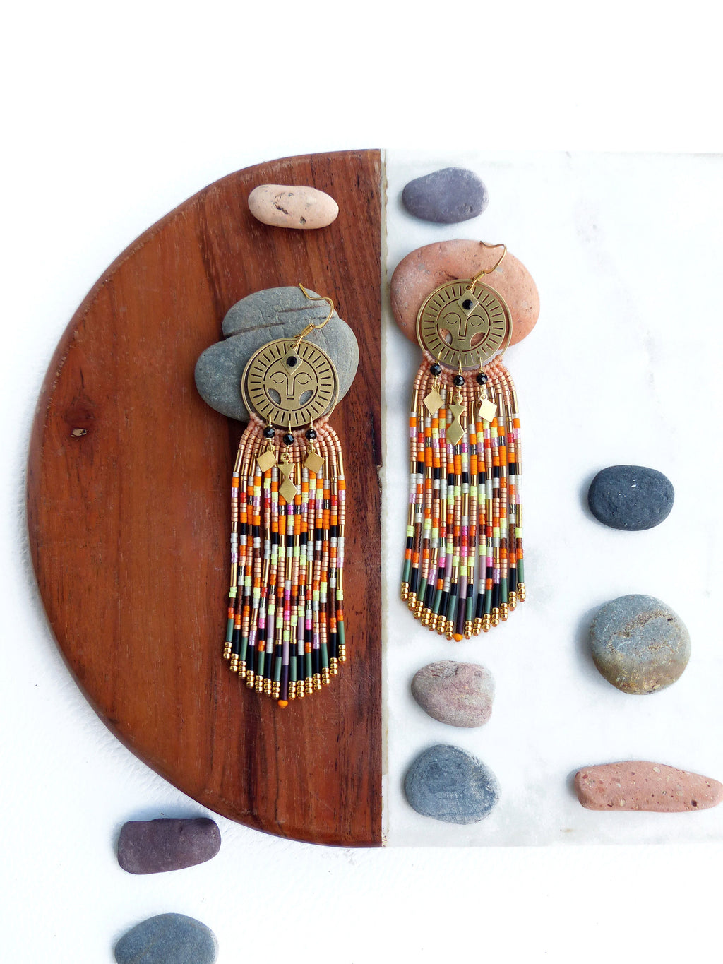 Peut inclure: Une paire de longues boucles d'oreilles en perles avec un motif de visage doré. Les boucles d'oreilles sont faites avec une variété de perles colorées, notamment orange, rose, vert et noir. Les boucles d'oreilles sont suspendues sur une surface en bois et en marbre.