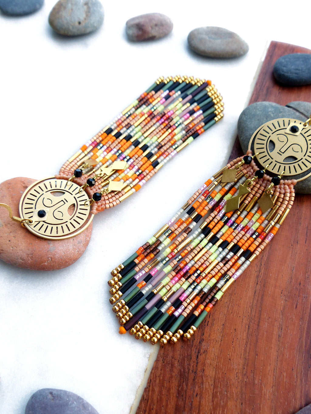 Peut inclure: Une paire de longues boucles d'oreilles en perles colorées avec des breloques circulaires dorées représentant un visage stylisé. Les boucles d'oreilles sont faites avec une variété de perles dans des tons d'orange, de rose, de vert, de noir et d'or.