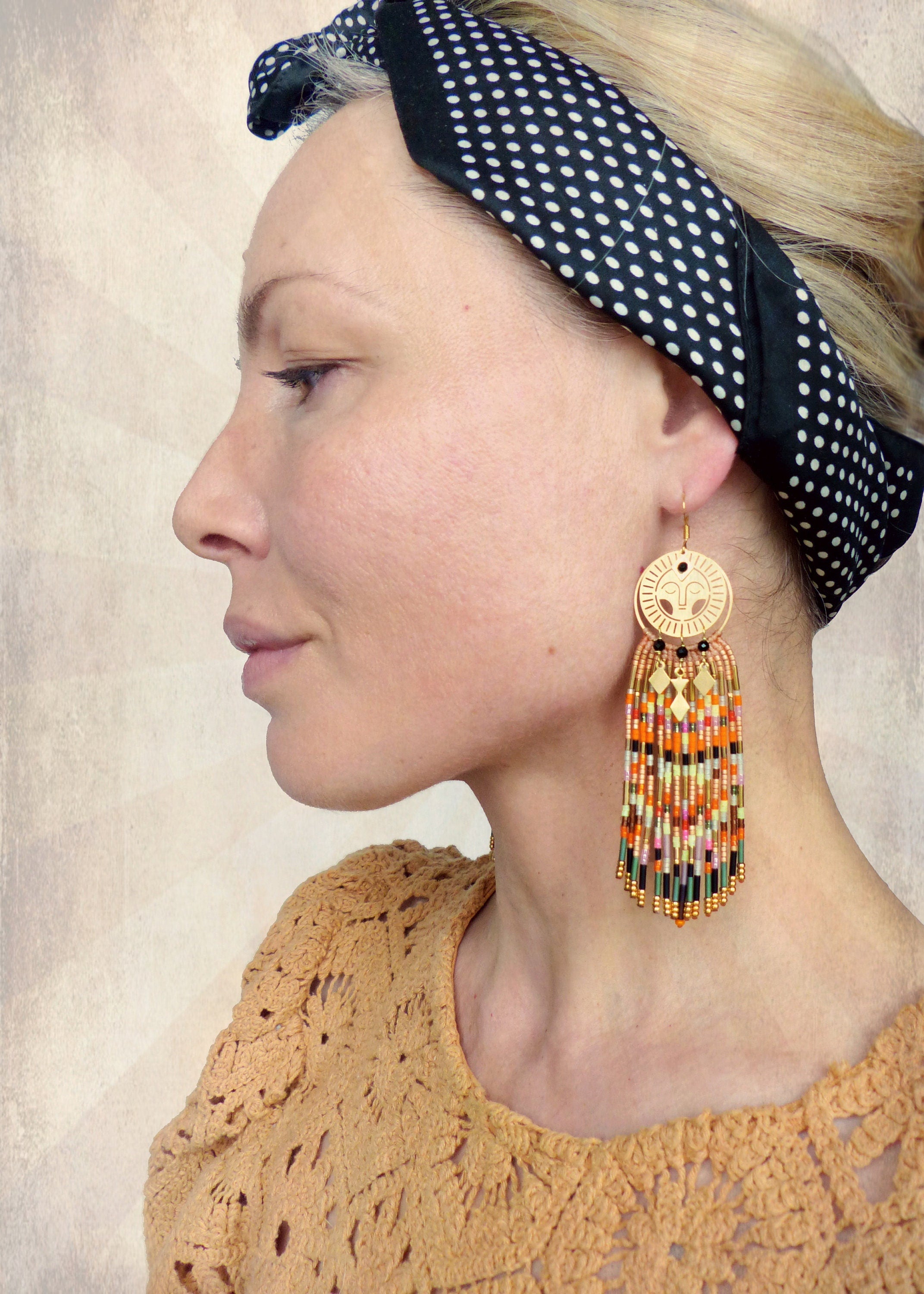 Peut inclure: Boucles d'oreilles dorées avec une frange de perles et un charme en forme de visage de soleil. Les boucles d'oreilles sont portées par une personne avec un bandeau à pois noirs et blancs.