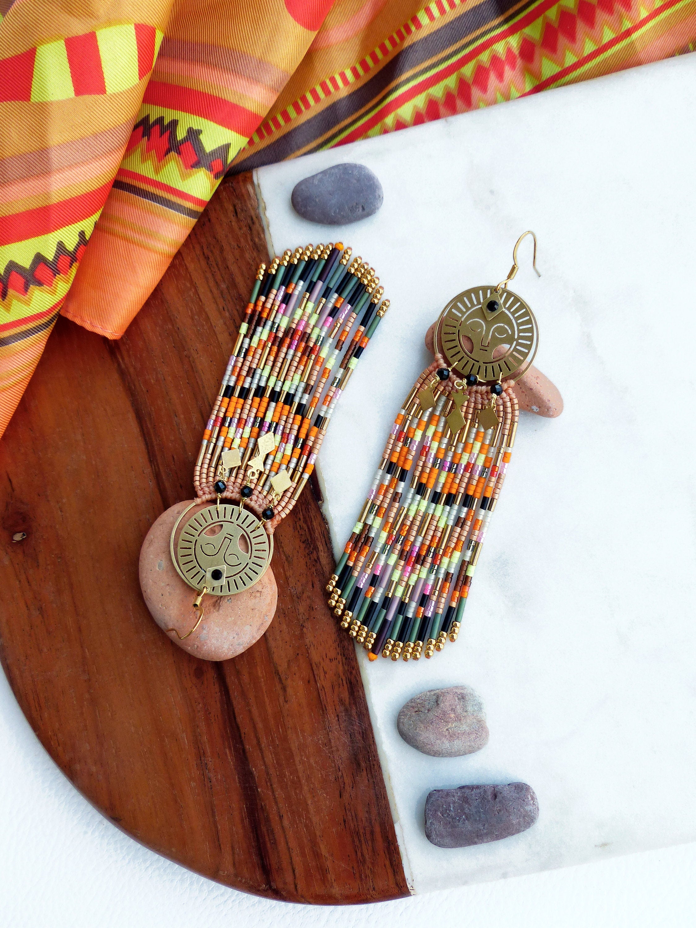 Peut inclure: Une paire de longues boucles d'oreilles en perles avec des breloques en forme de soleil dorés. Les boucles d'oreilles sont faites avec une variété de perles colorées, notamment noires, orange, vertes et roses. Les boucles d'oreilles sont suspendues sur une surface en bois avec un fond blanc.