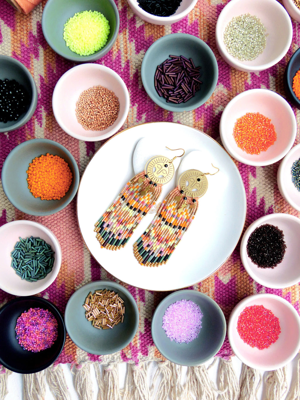 Peut inclure: Une paire de boucles d'oreilles en perles dorées et multicolores avec un pendentif circulaire. Les boucles d'oreilles sont présentées sur une assiette blanche avec de petits bols de perles de différentes couleurs entourant l'assiette.