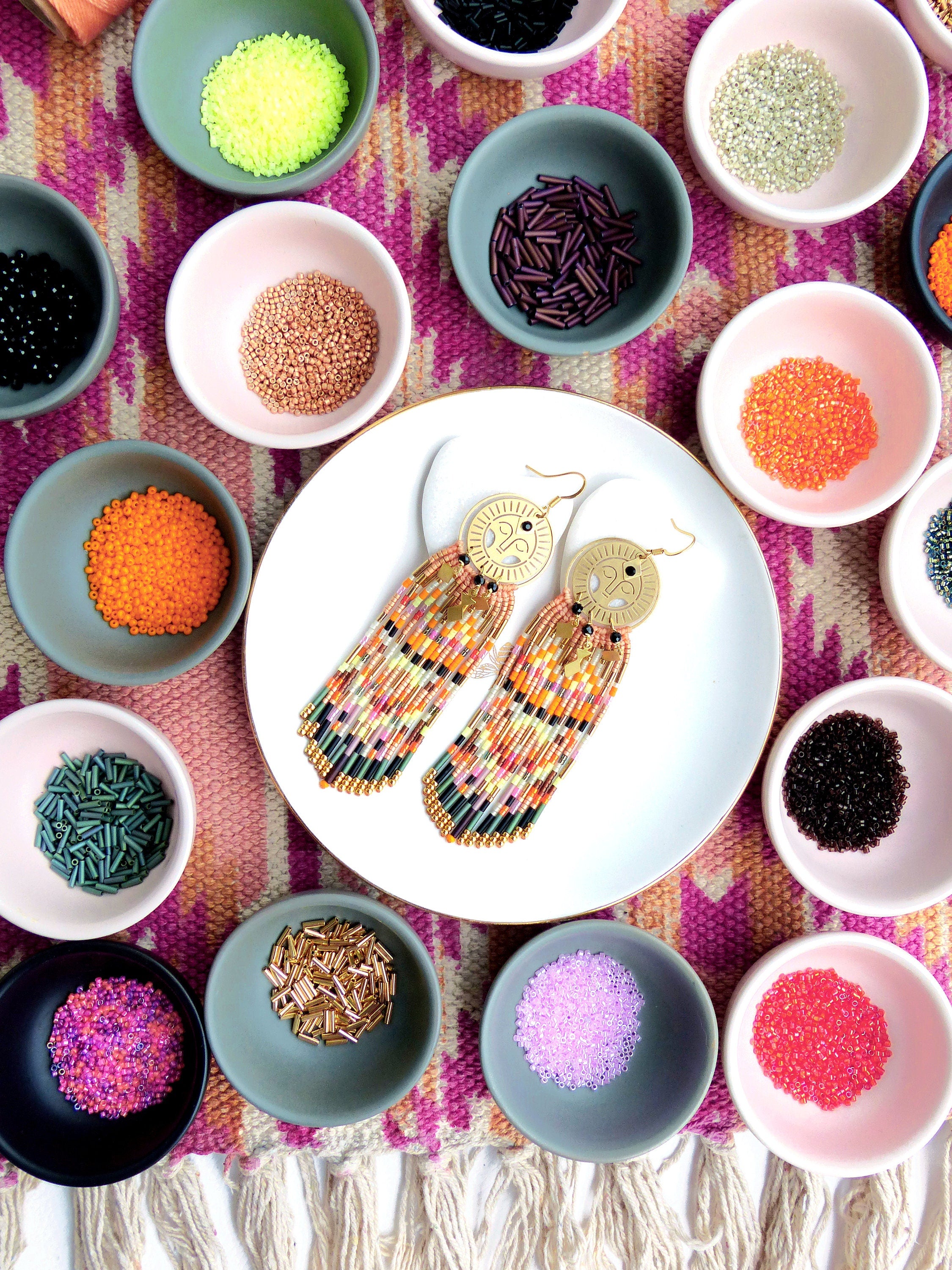 Peut inclure: Une paire de boucles d'oreilles en perles dorées et multicolores avec un pendentif circulaire. Les boucles d'oreilles sont présentées sur une assiette blanche avec de petits bols de perles de différentes couleurs entourant l'assiette.