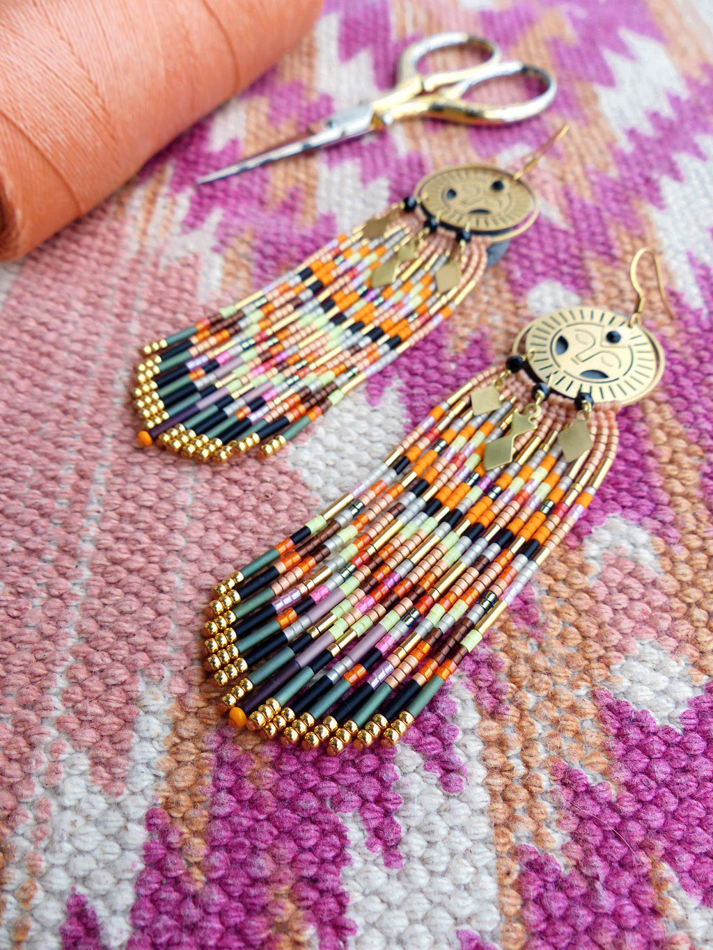 Peut inclure: Une paire de longues boucles d'oreilles en perles avec des accents dorés. Les boucles d'oreilles présentent un charme circulaire doré avec un motif géométrique. Les perles sont dans des tons d'orange, de rose, de vert et de noir.