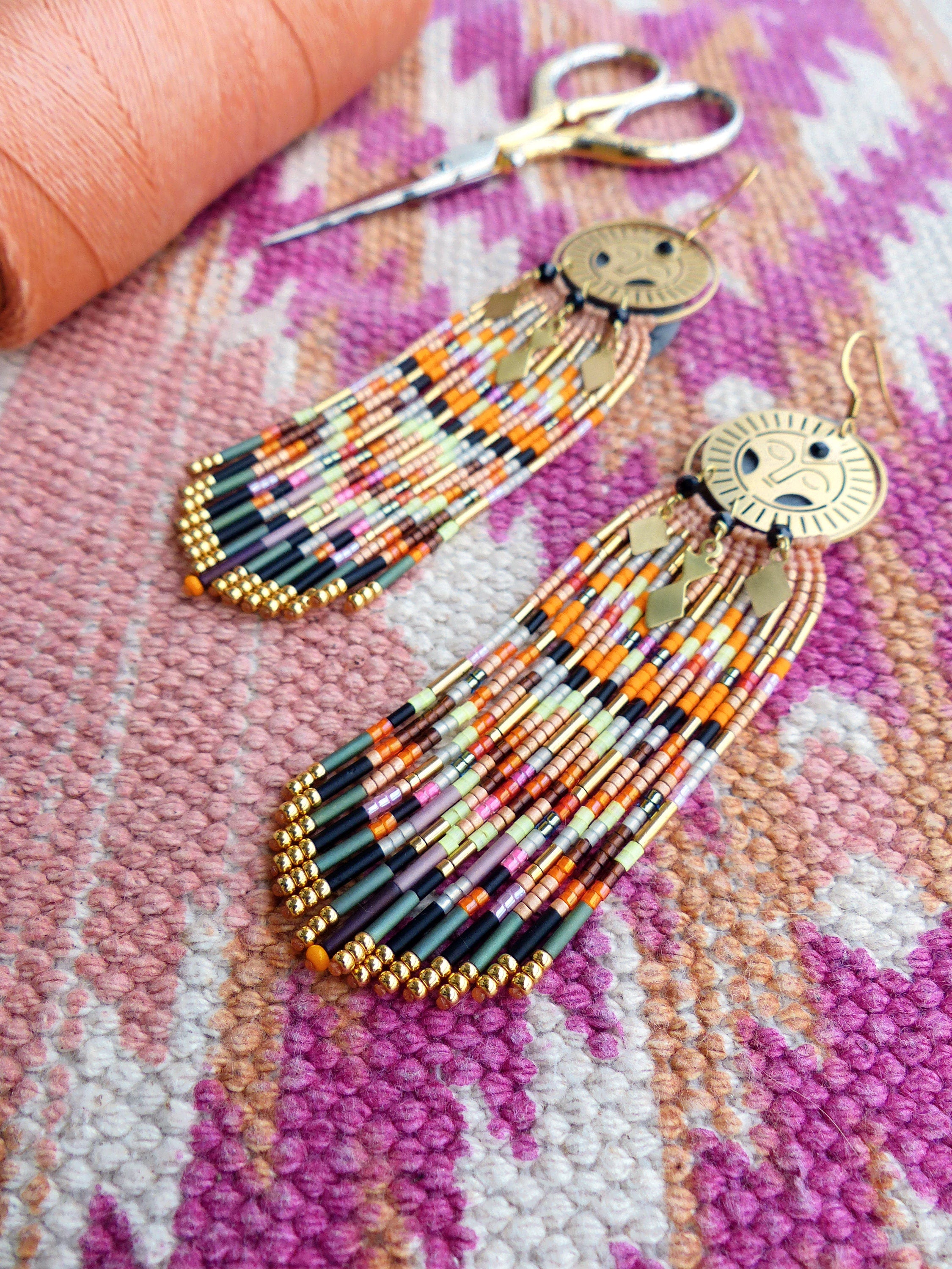 Peut inclure: Une paire de longues boucles d'oreilles en perles avec des accents dorés. Les boucles d'oreilles présentent un charme circulaire doré avec un motif géométrique. Les perles sont dans des tons d'orange, de rose, de vert et de noir.