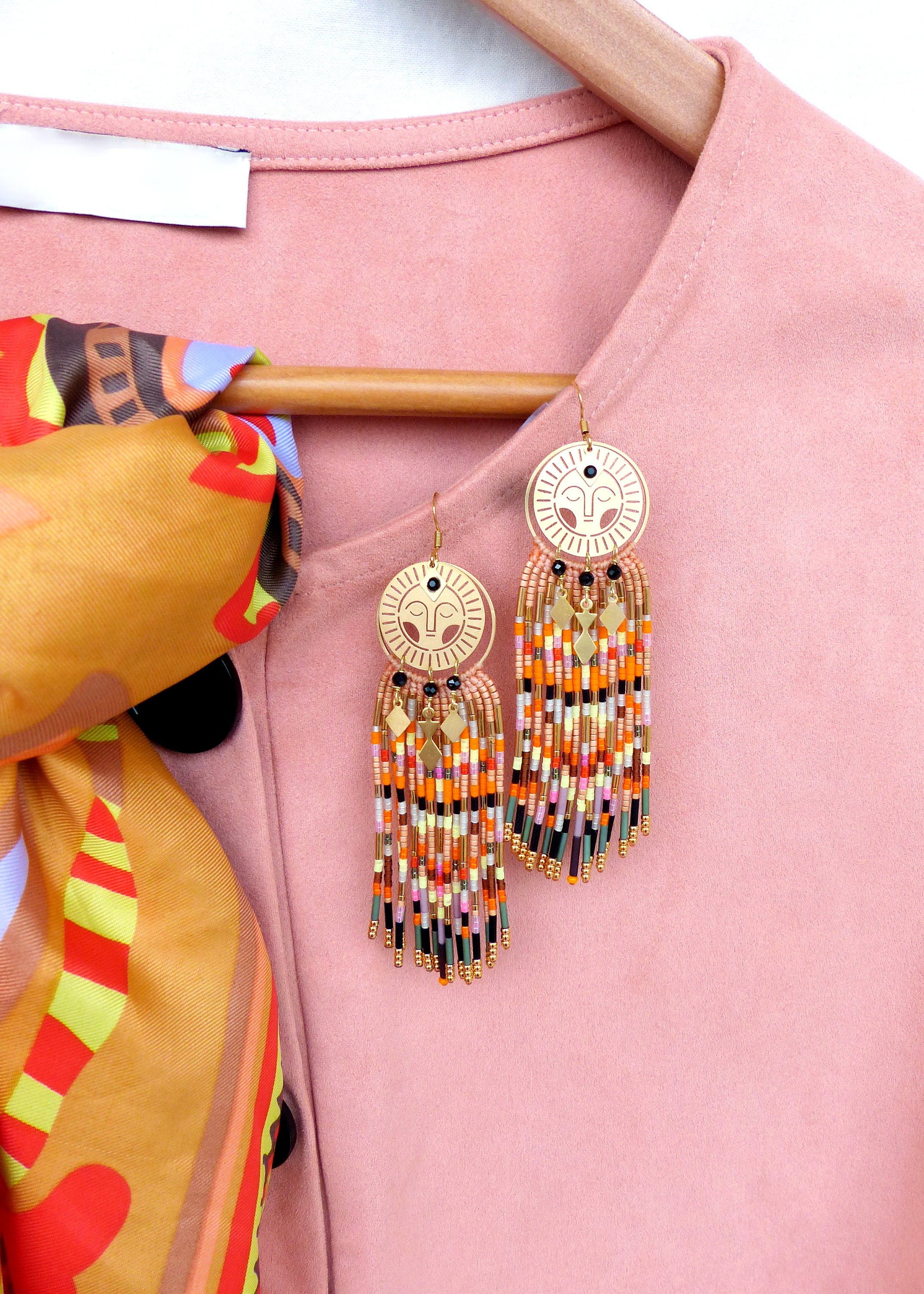 Peut inclure: Une paire de boucles d'oreilles dorées avec un motif de visage et une frange de perles colorées. Les boucles d'oreilles sont accrochées à un cintre rose.