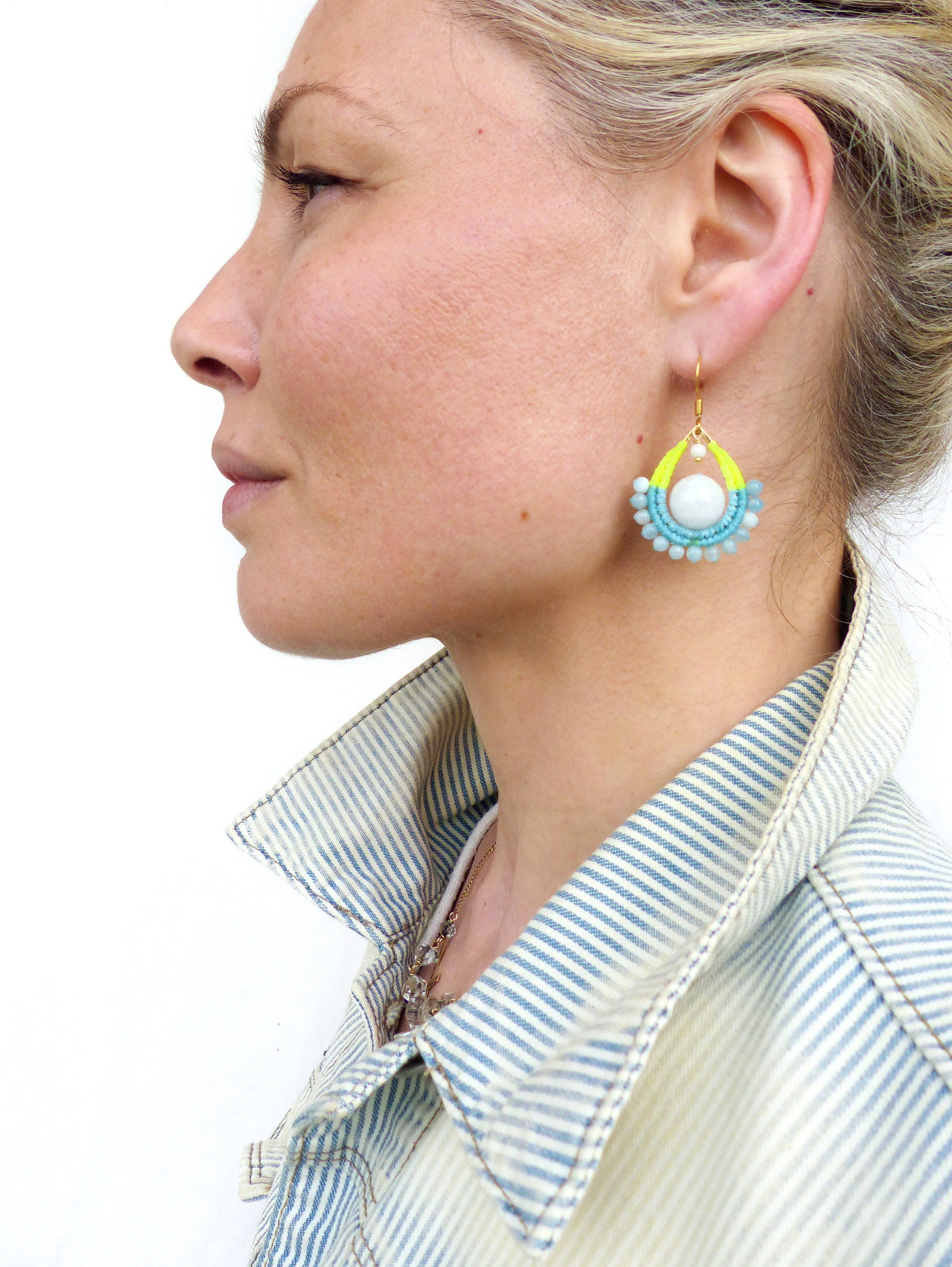 Peut inclure: Une paire de boucles d'oreilles en forme de goutte d'eau avec une perle blanche au centre. Les boucles d'oreilles sont faites de perles turquoise et jaunes et ont un crochet en or.