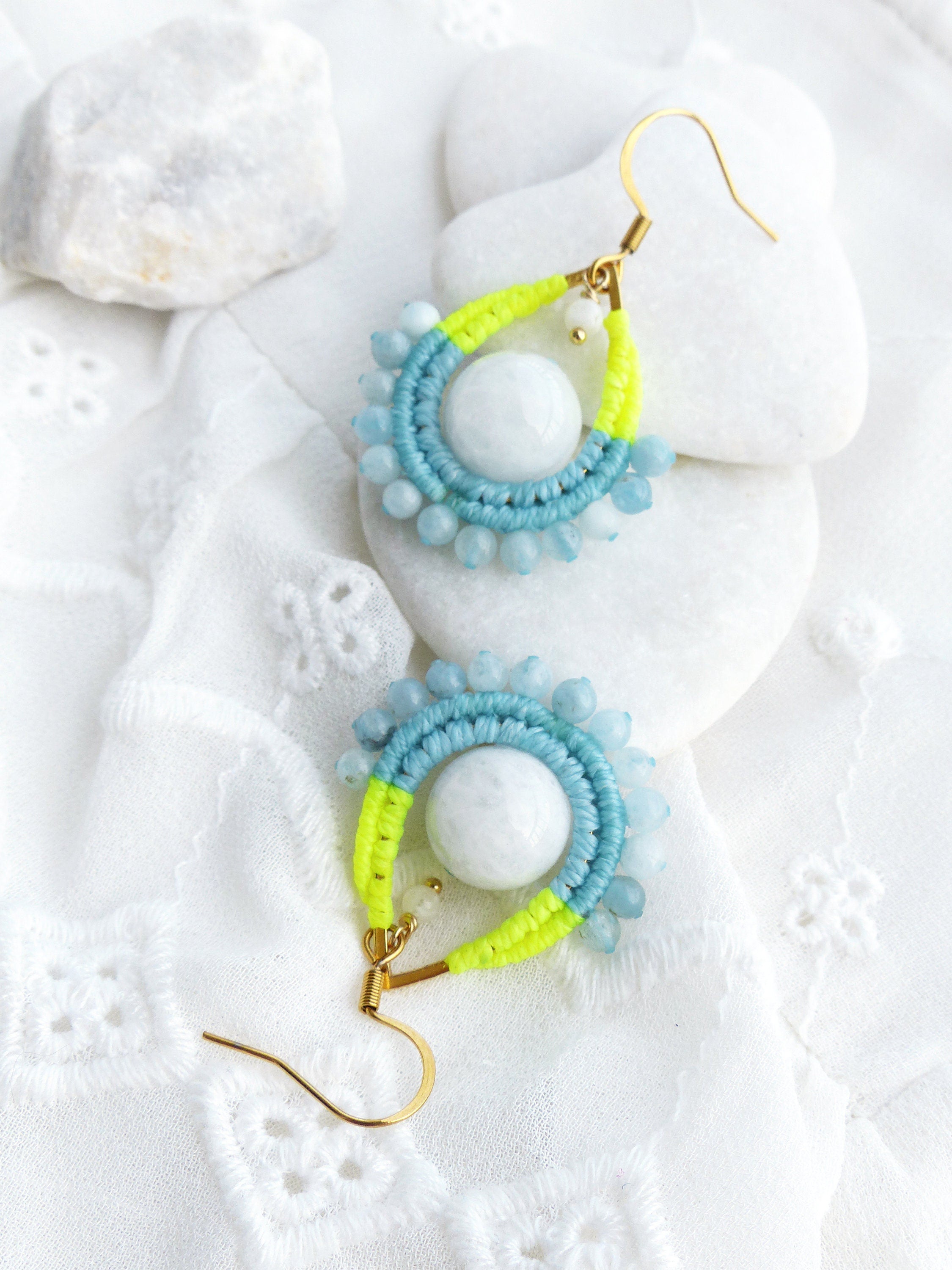 Peut inclure: Une paire de boucles d'oreilles pendantes avec un design en macramé bleu clair et jaune fluo. Les boucles d'oreilles présentent une pierre blanche au centre de chaque design.