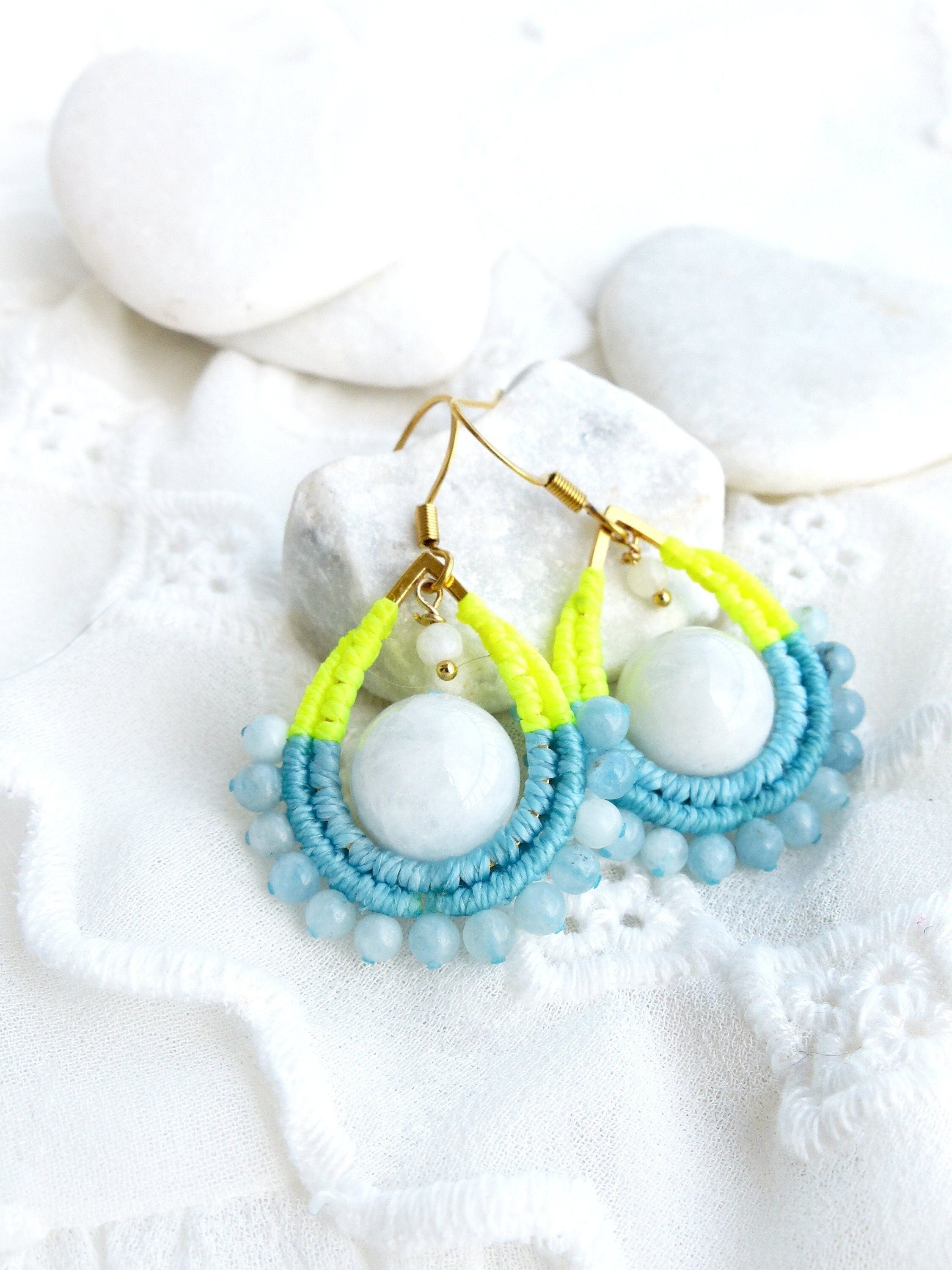 Peut inclure: Une paire de boucles d'oreilles pendantes avec une pierre bleu clair et blanche, entourée de petites perles bleu clair. Les boucles d'oreilles sont faites avec de la ficelle jaune fluo et bleu clair.