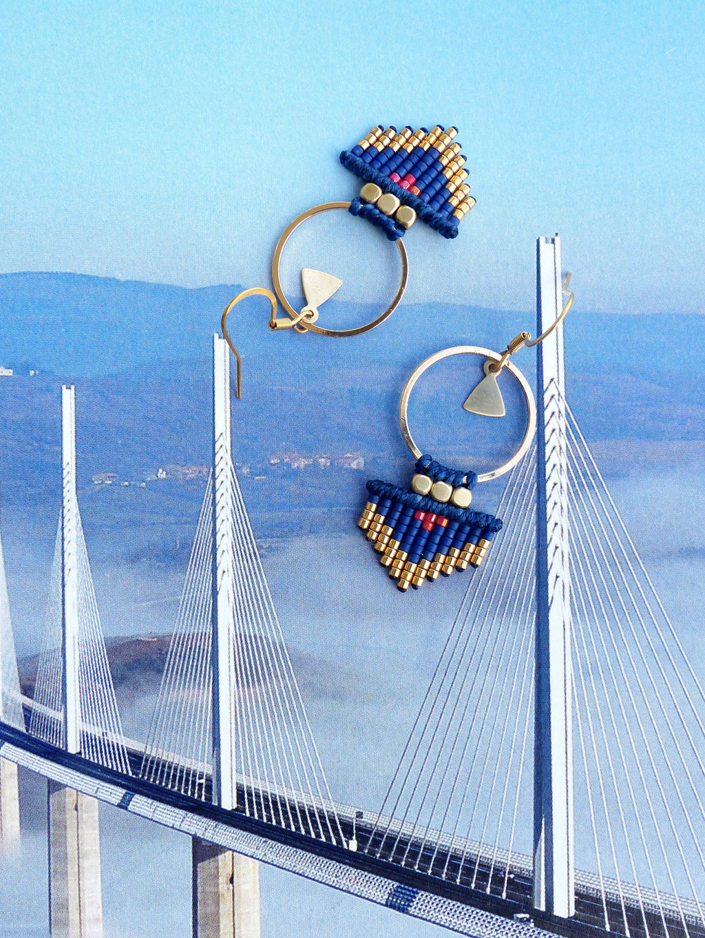 Peut inclure: Une paire de boucles d'oreilles créoles dorées avec des franges de perles bleues et dorées et des accents rouges. Les boucles d'oreilles sont suspendues sur un fond de pont.