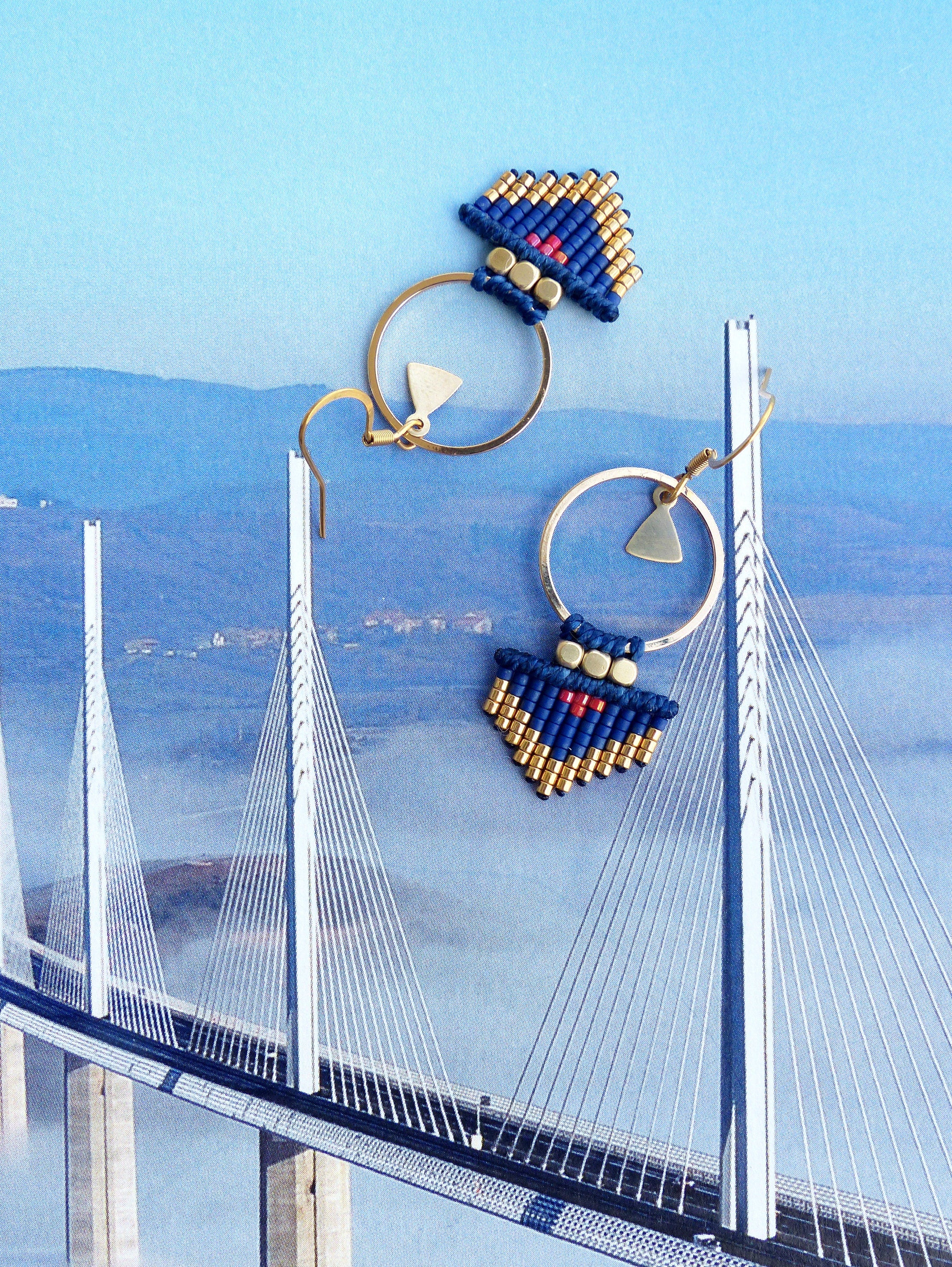 Peut inclure: Une paire de boucles d'oreilles créoles dorées avec des franges de perles bleues et dorées et des accents rouges. Les boucles d'oreilles sont suspendues sur un fond de pont.