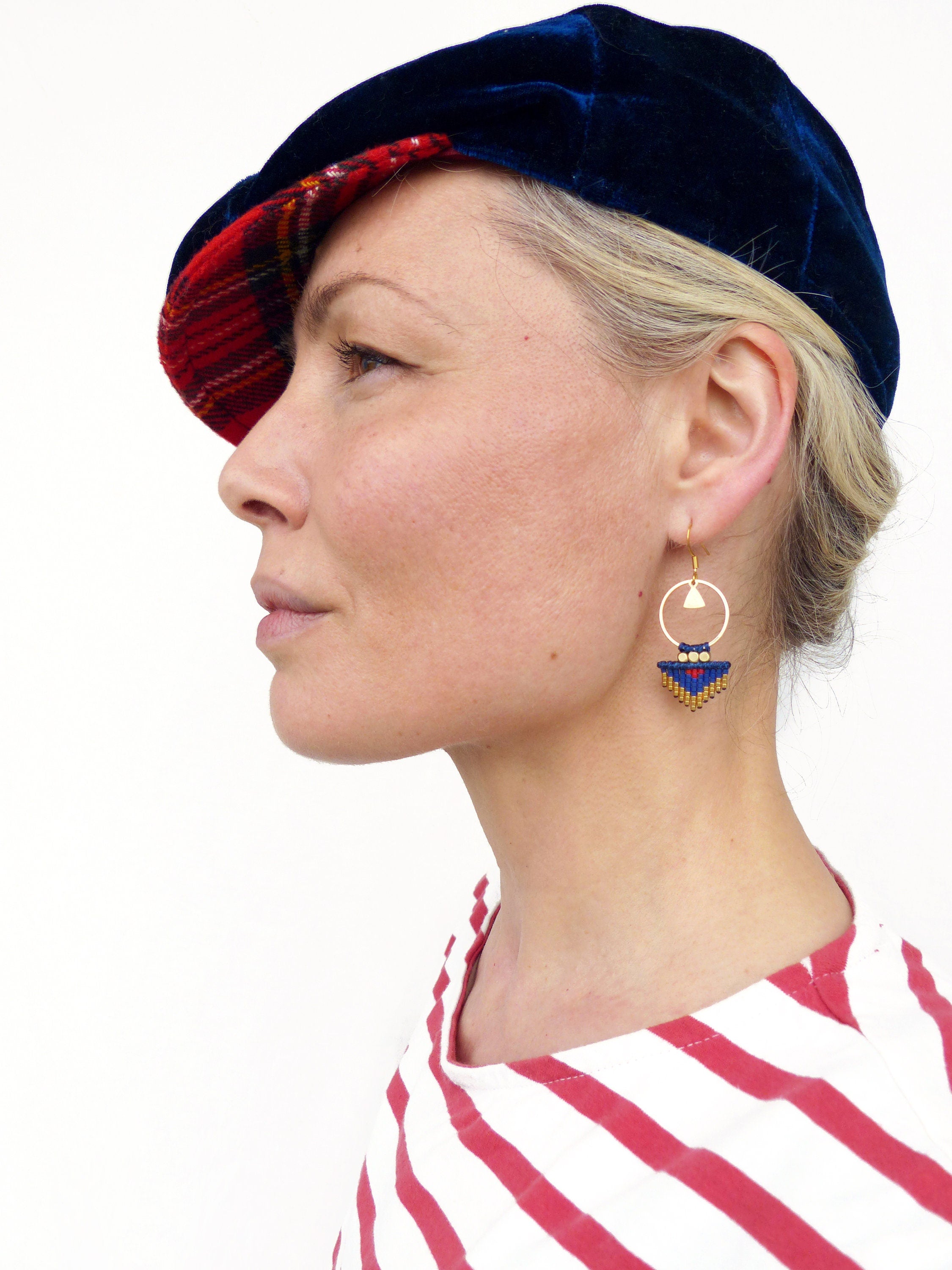 Peut inclure: Une femme portant une casquette de newsboy en velours bleu et en tartan rouge avec des boucles d'oreilles créoles dorées ornées de breloques en perles bleues et dorées. Elle porte une chemise à rayures rouges et blanches.